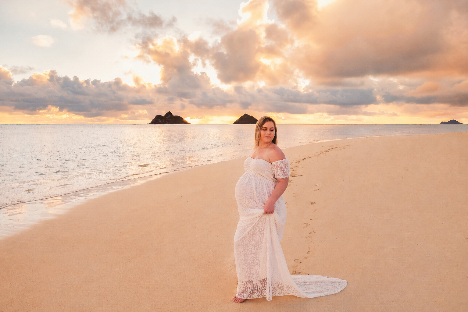 Pregnant woman in a white lace dress standing barefoot on a sandy beach during sunset, with clouds and distant islands in the background.