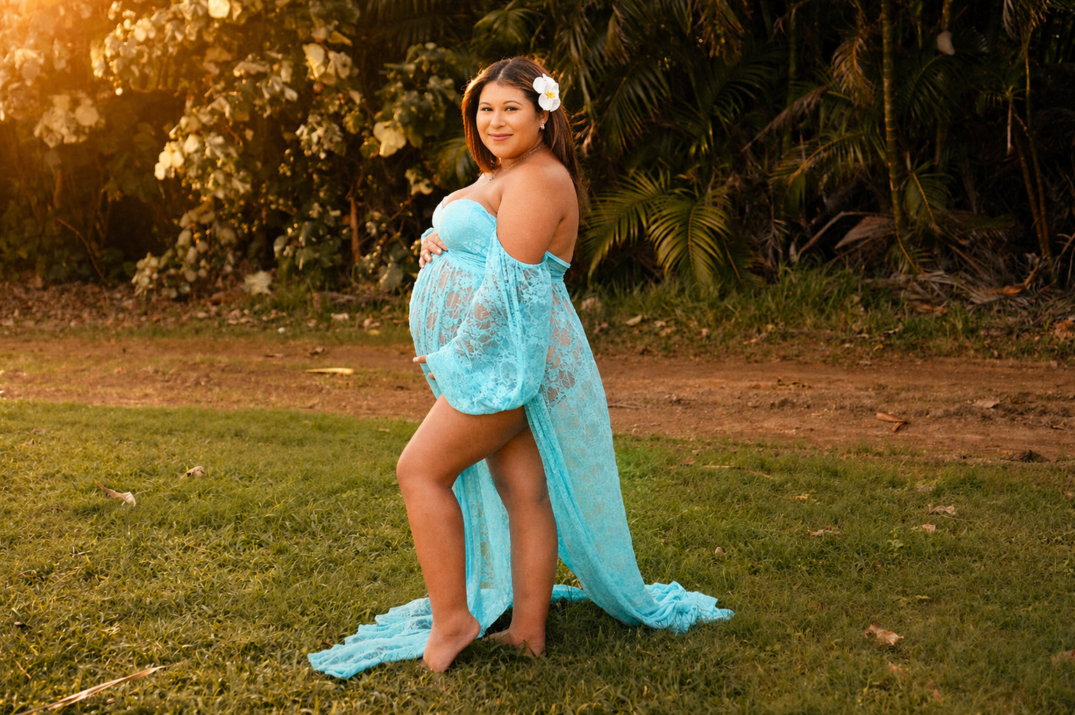 A pregnant woman in a light blue, lace dress with off-the-shoulder sleeves standing outdoors, smiling and holding her pregnant belly, with a flower in her hair and lush greenery in the background during sunset.