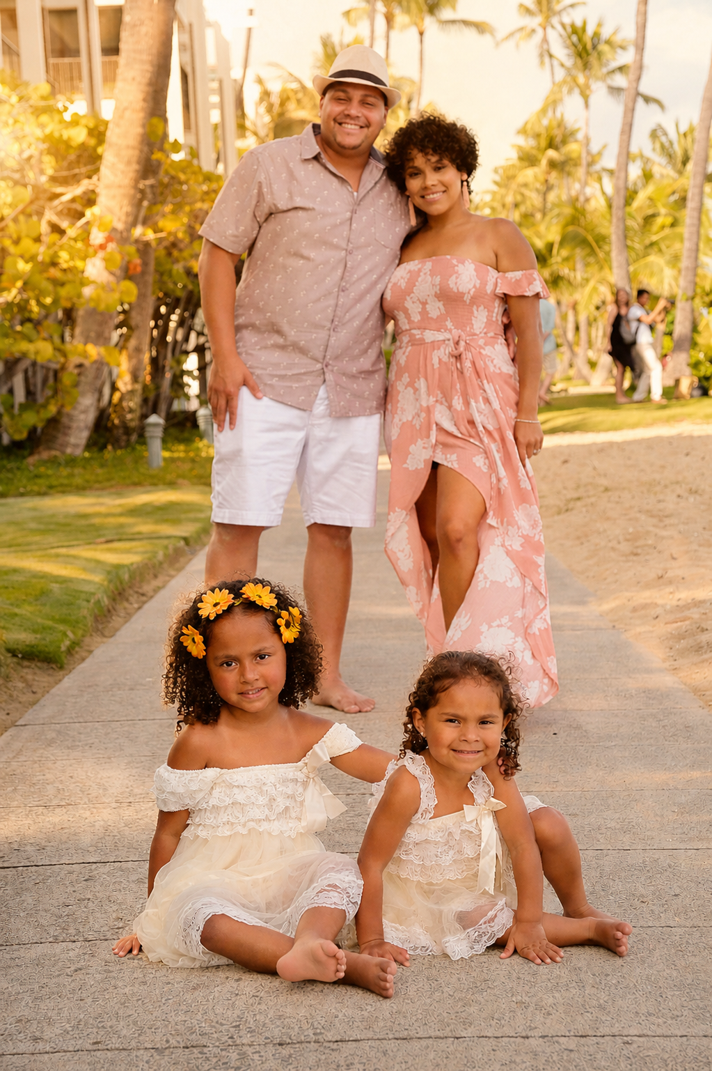 A family of five smiling on a tropical beach sidewalk with palm trees. Two young girls sit on the ground in front, wearing cream-colored dresses; one has a yellow flower headband. Two adults, a man and a woman, stand behind them, smiling. The man wea