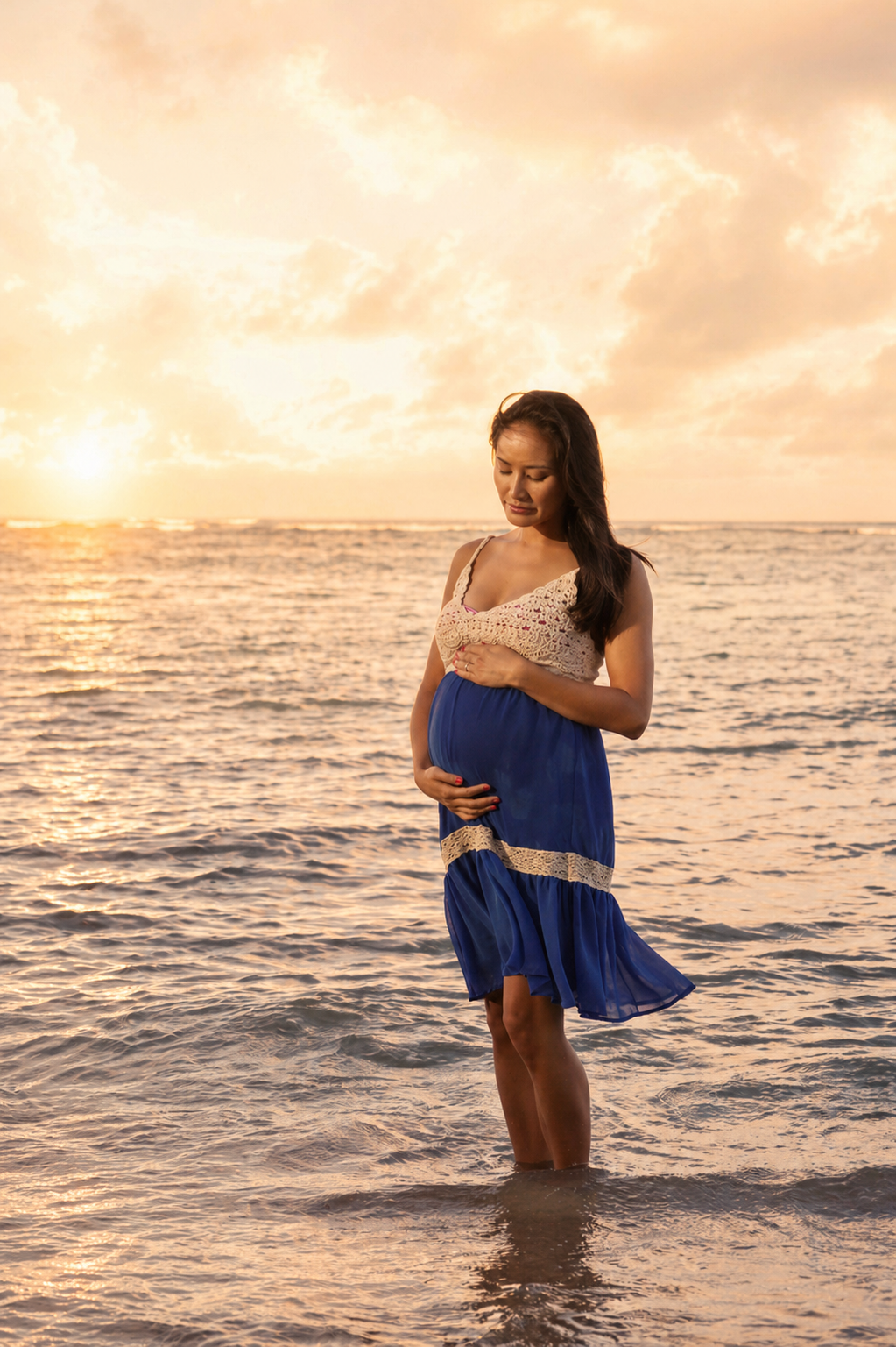 A pregnant woman stands in the ocean at sunset, holding her belly with one hand and her chest with the other, wearing a blue dress with lace details.