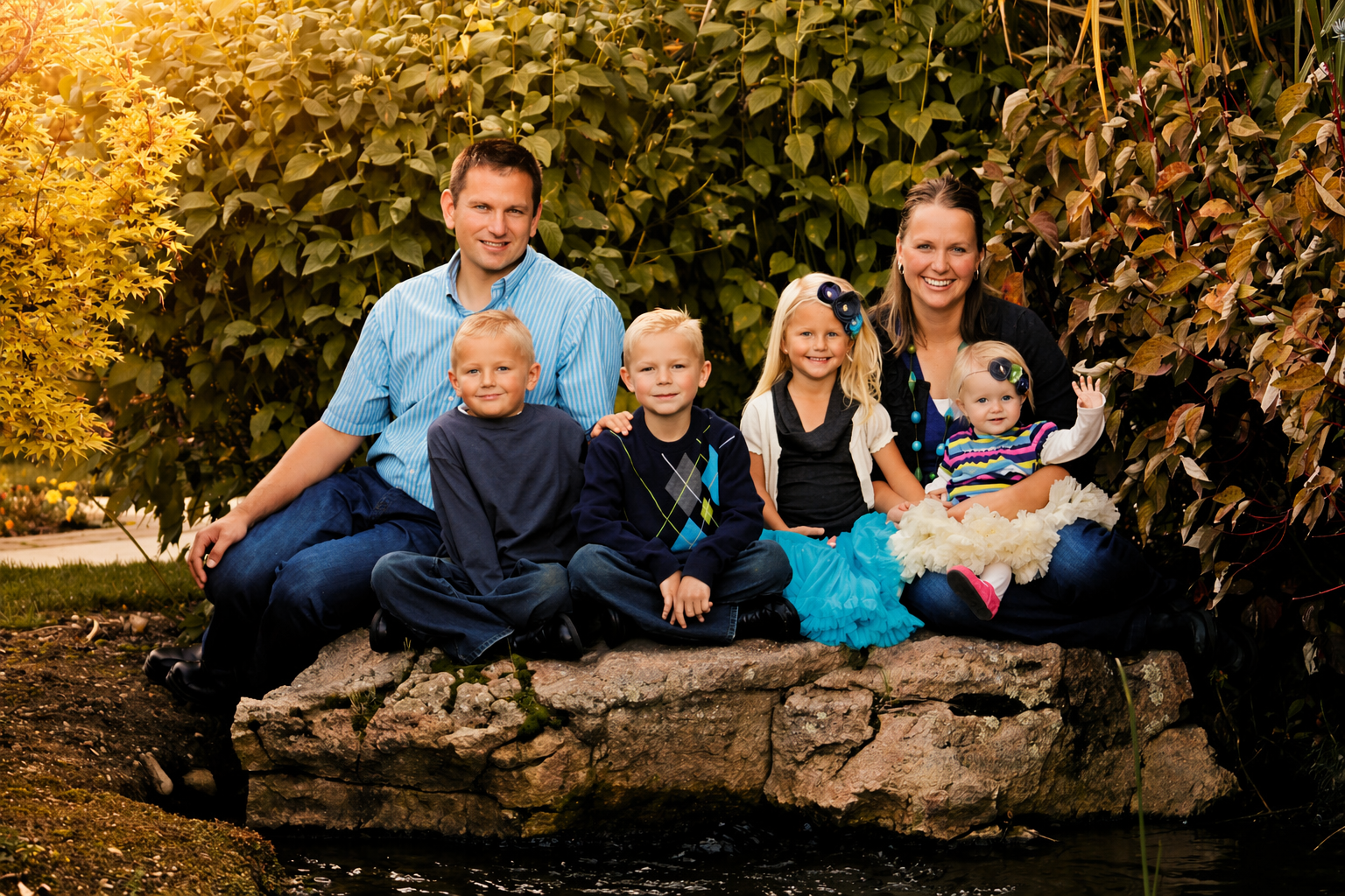 Family of seven sitting on a rock by water, surrounded by lush greenery and plants, during daytime.