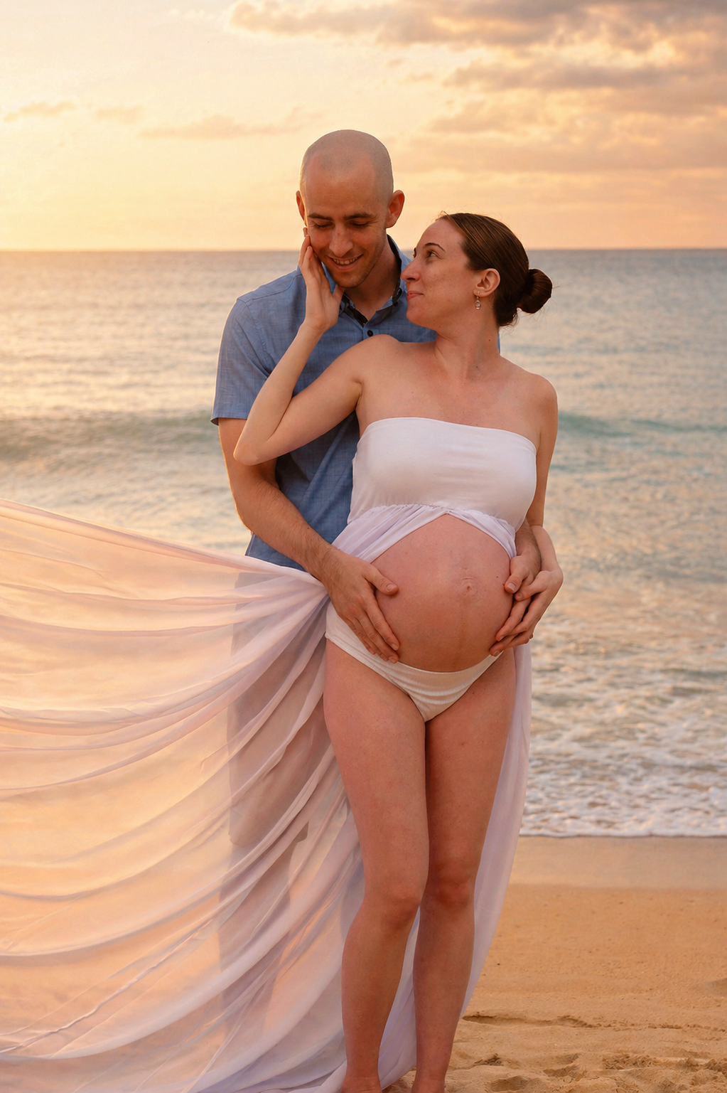 A couple on the beach at sunset, the woman is pregnant and wearing a flowing white dress, the man is behind her, touching her belly, and they are smiling at each other.