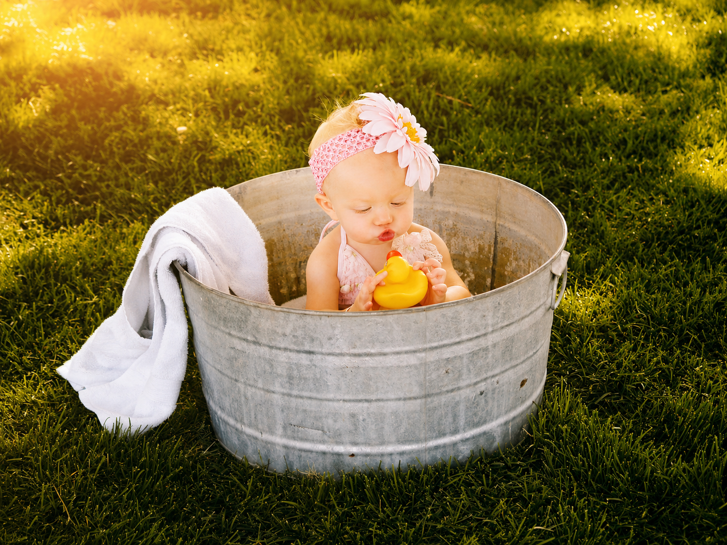 A baby girl with a pink headband and large flower sitting in a metal tub outdoors on grass, holding a yellow rubber duck, with a white towel hanging over the tub's side.