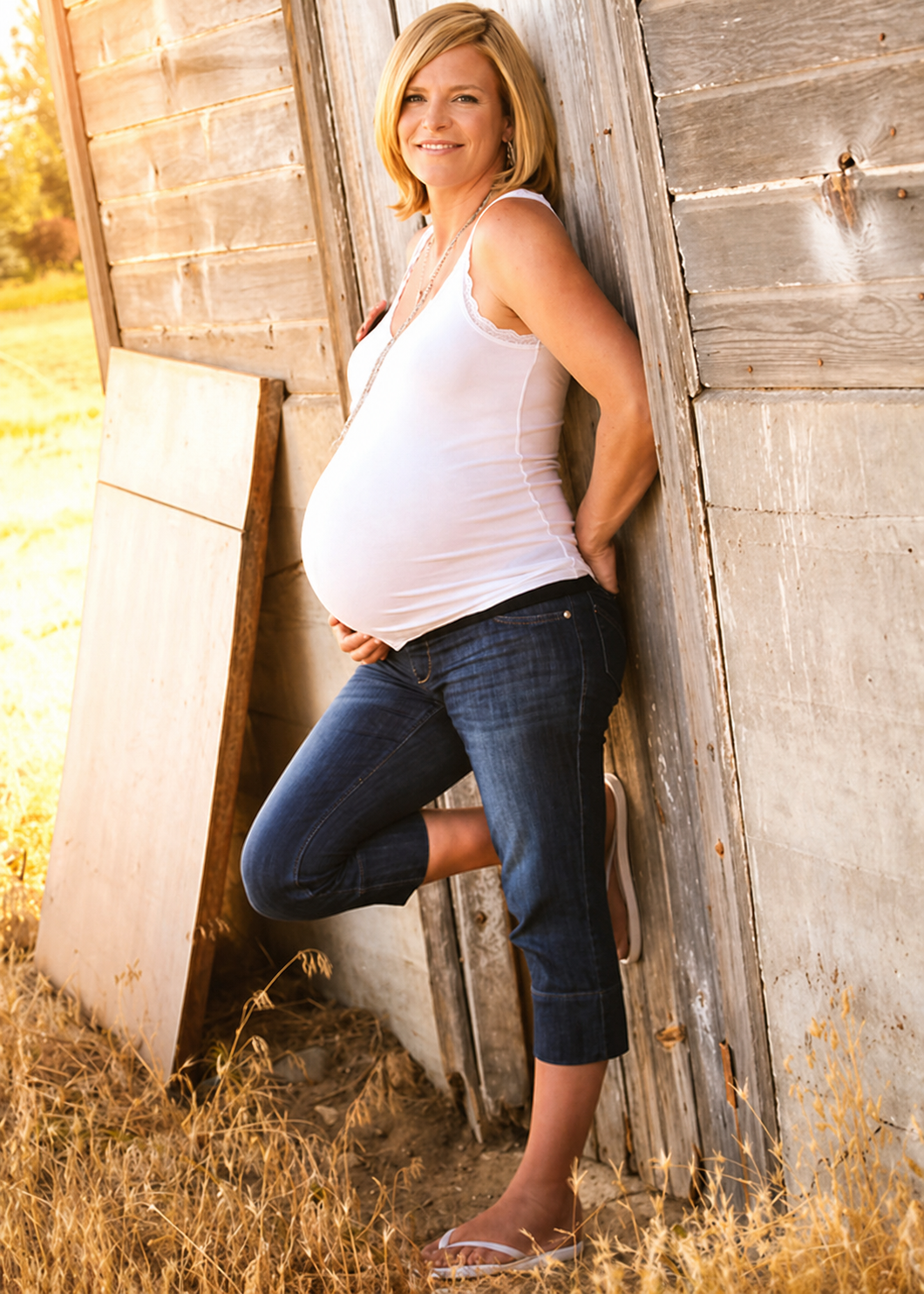 Pregnant woman with blonde hair leaning against wooden shed outside, wearing a white tank top, jeans, and flip-flops, smiling at the camera.