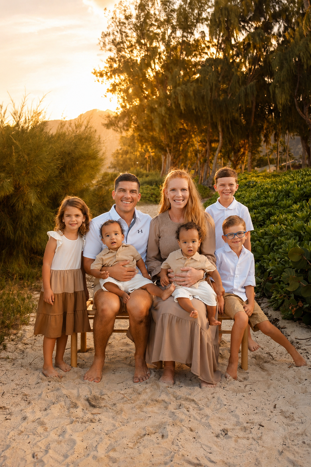 A family of nine sitting on a wooden bench on a sandy beach during sunset, with trees and hills in the background.