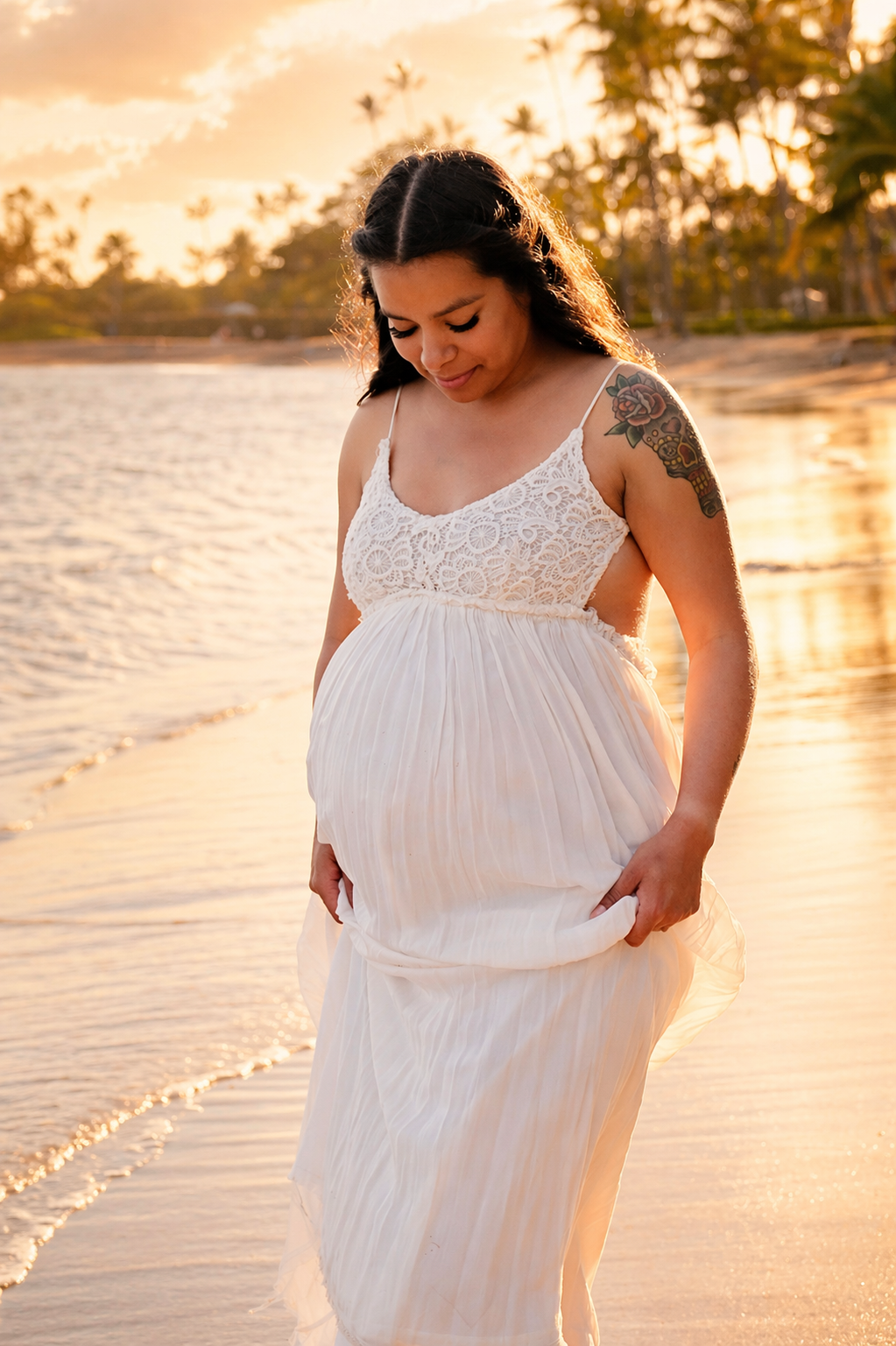 A pregnant woman in a white lace and chiffon dress stands on the beach during sunset, gently holding her flowing dress and looking down.