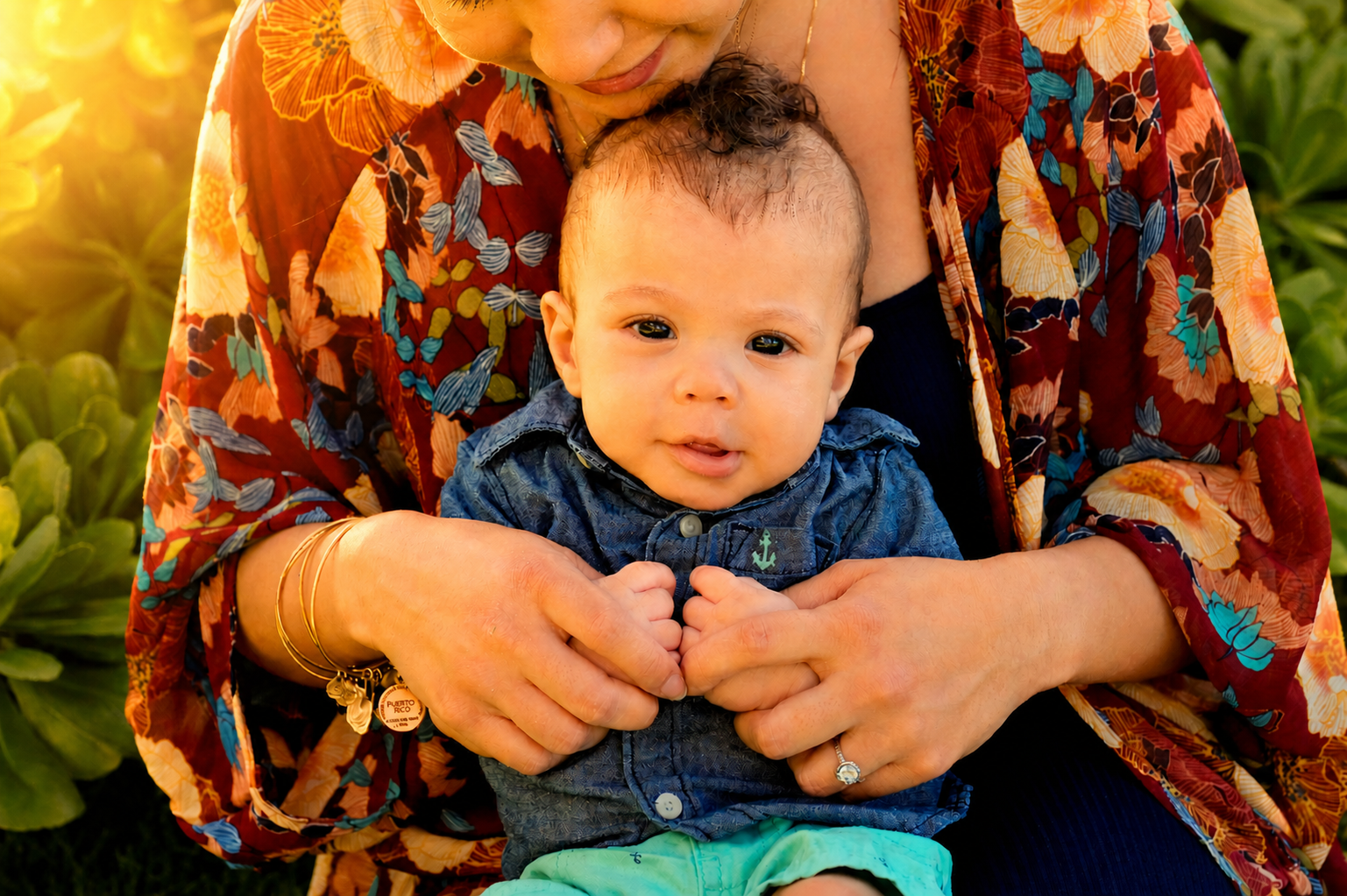 A baby boy with light skin and curly hair being held by an adult woman with tan skin and wearing a floral red and orange kimono. The baby is looking at the camera and has a slight smile, wearing a blue shirt with an anchor embroidered on it. The woma