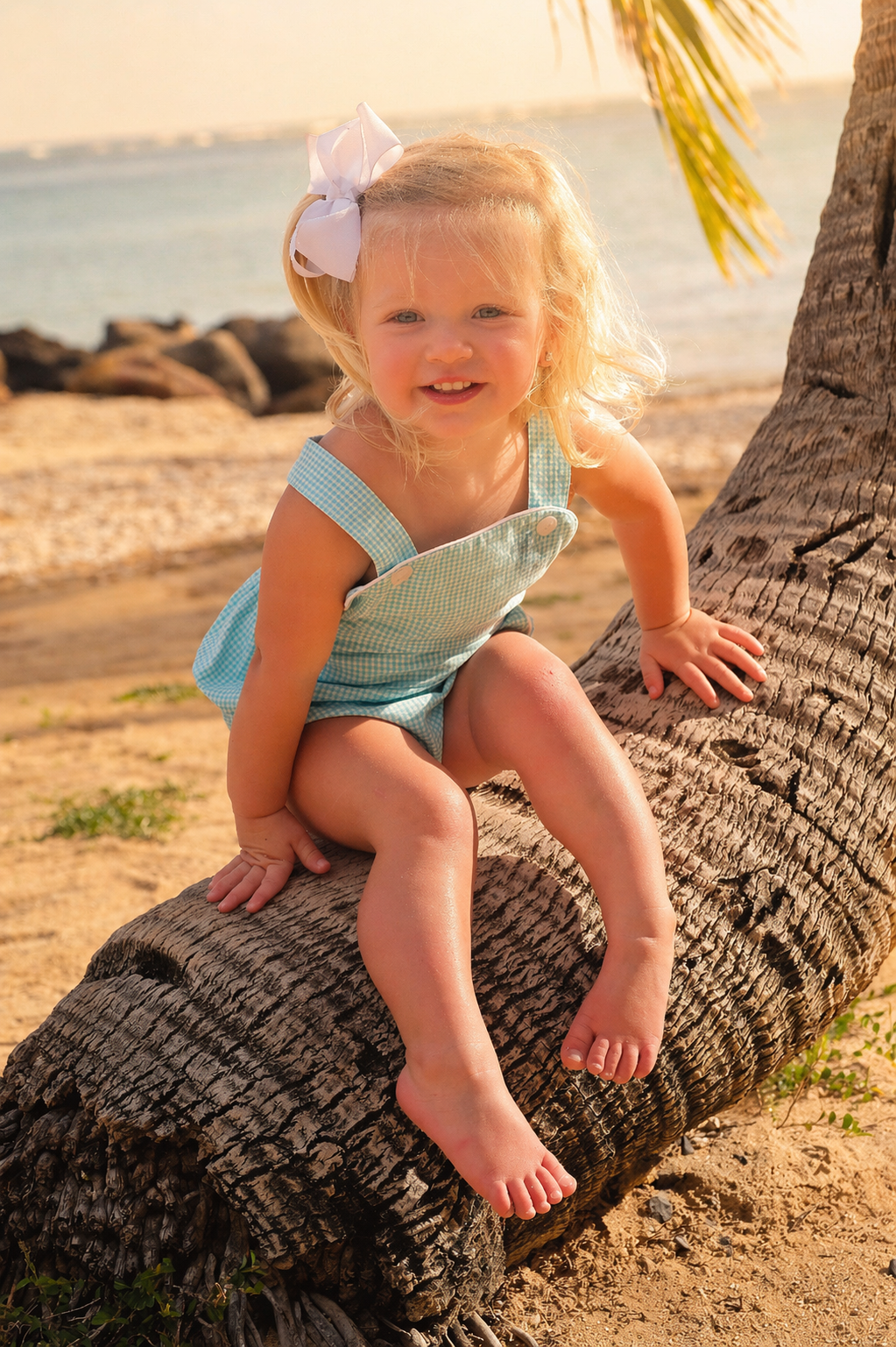A young girl with blonde hair, wearing a light blue checkered romper with a white bow in her hair, playfully climbing on a leaning palm tree on a sandy beach.