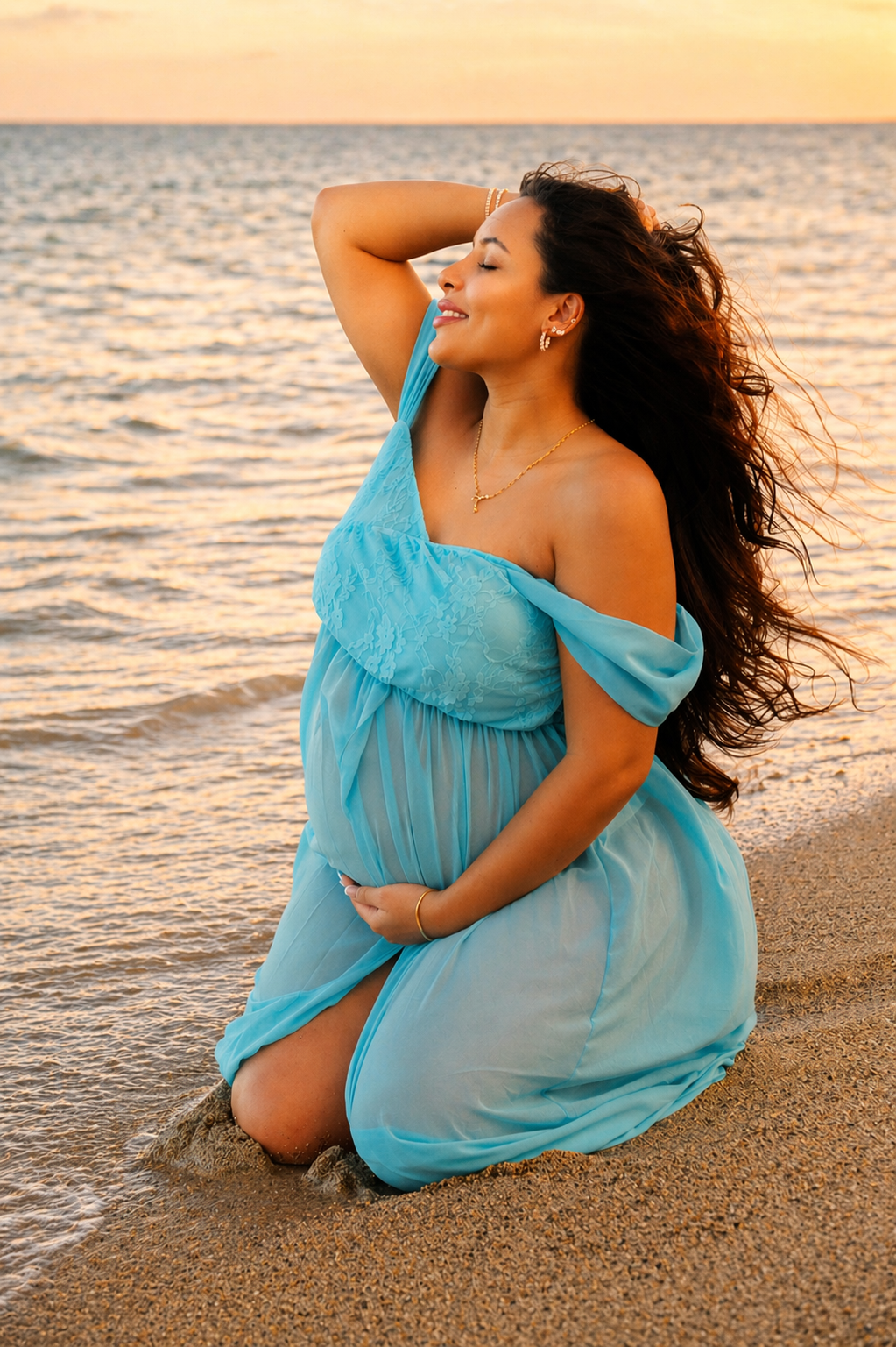 A pregnant woman in a blue dress kneeling on the beach at sunset, smiling with eyes closed, and holding her belly.