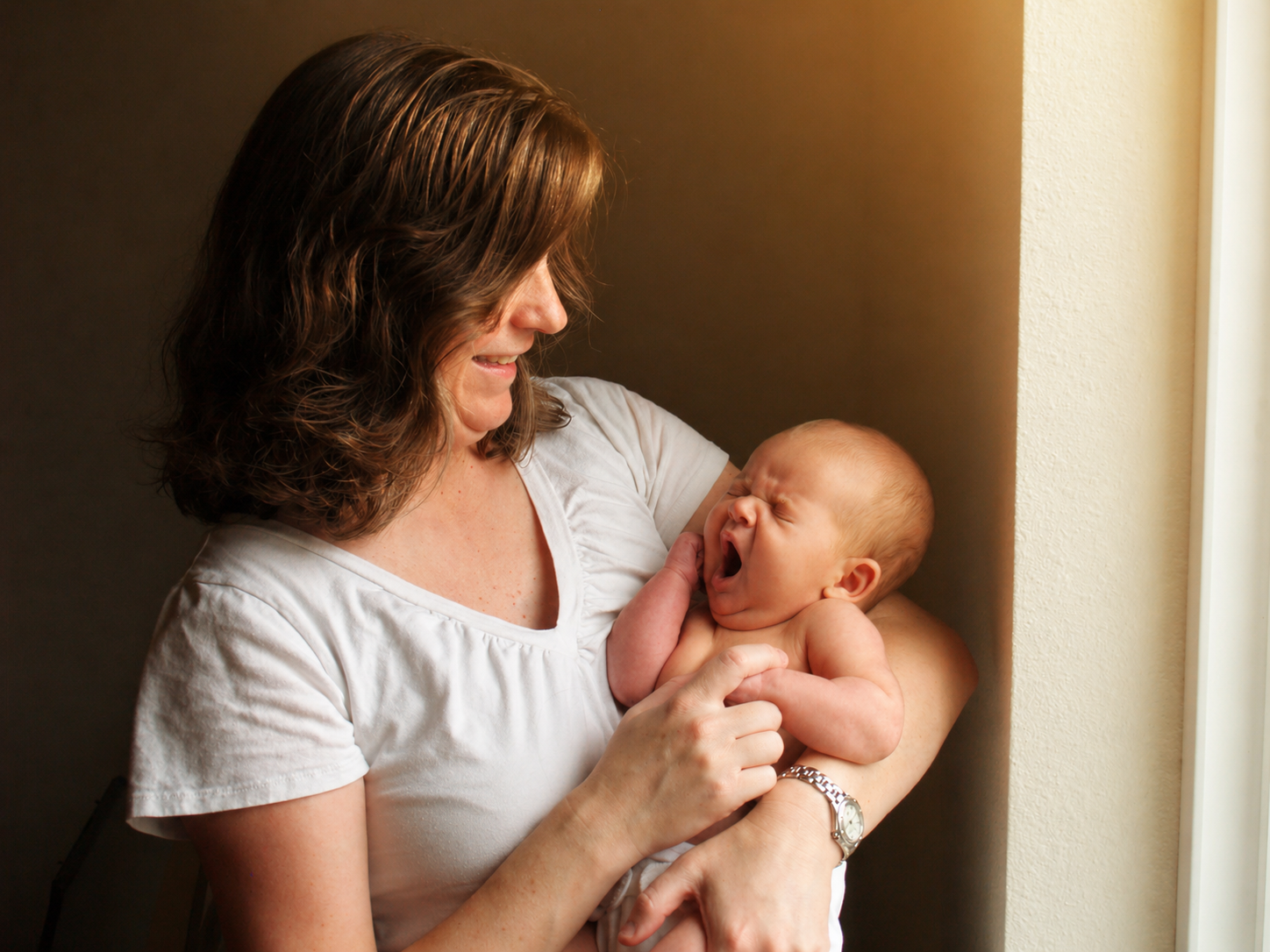 A woman holding a yawning baby near a window.