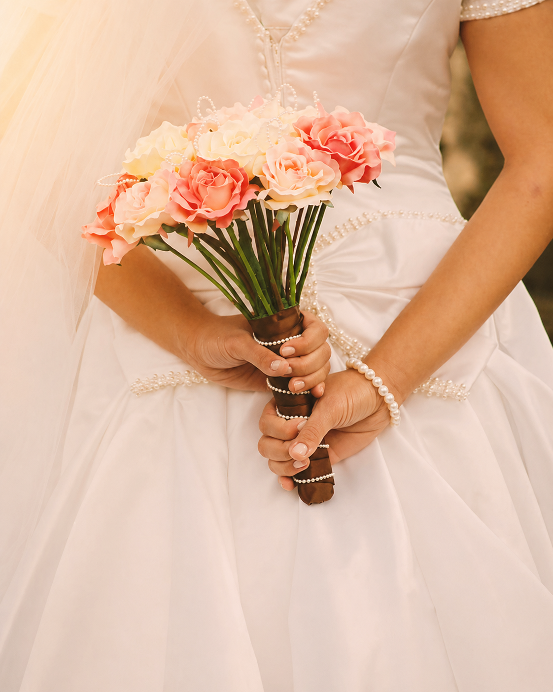 A bride holding a bouquet of pink and cream roses while wearing a white wedding gown.