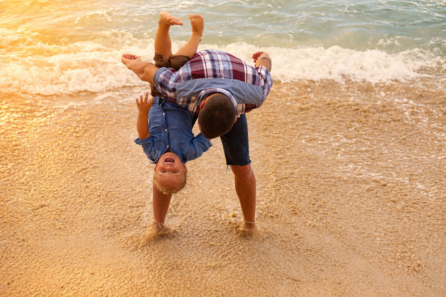 A man and a young boy playing and laughing on the beach, with the man holding the boy upside down over the sand and water as the sun sets in the background.