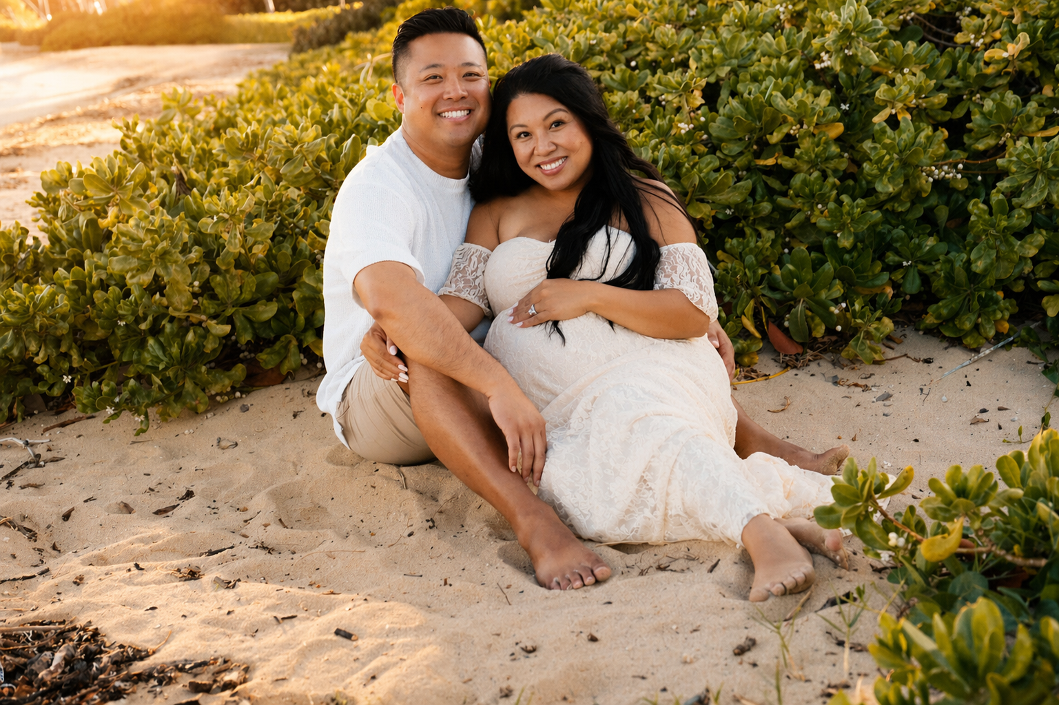 A smiling couple sitting on the sand near green bushes at sunset, with the woman pregnant and wearing a white lace dress.