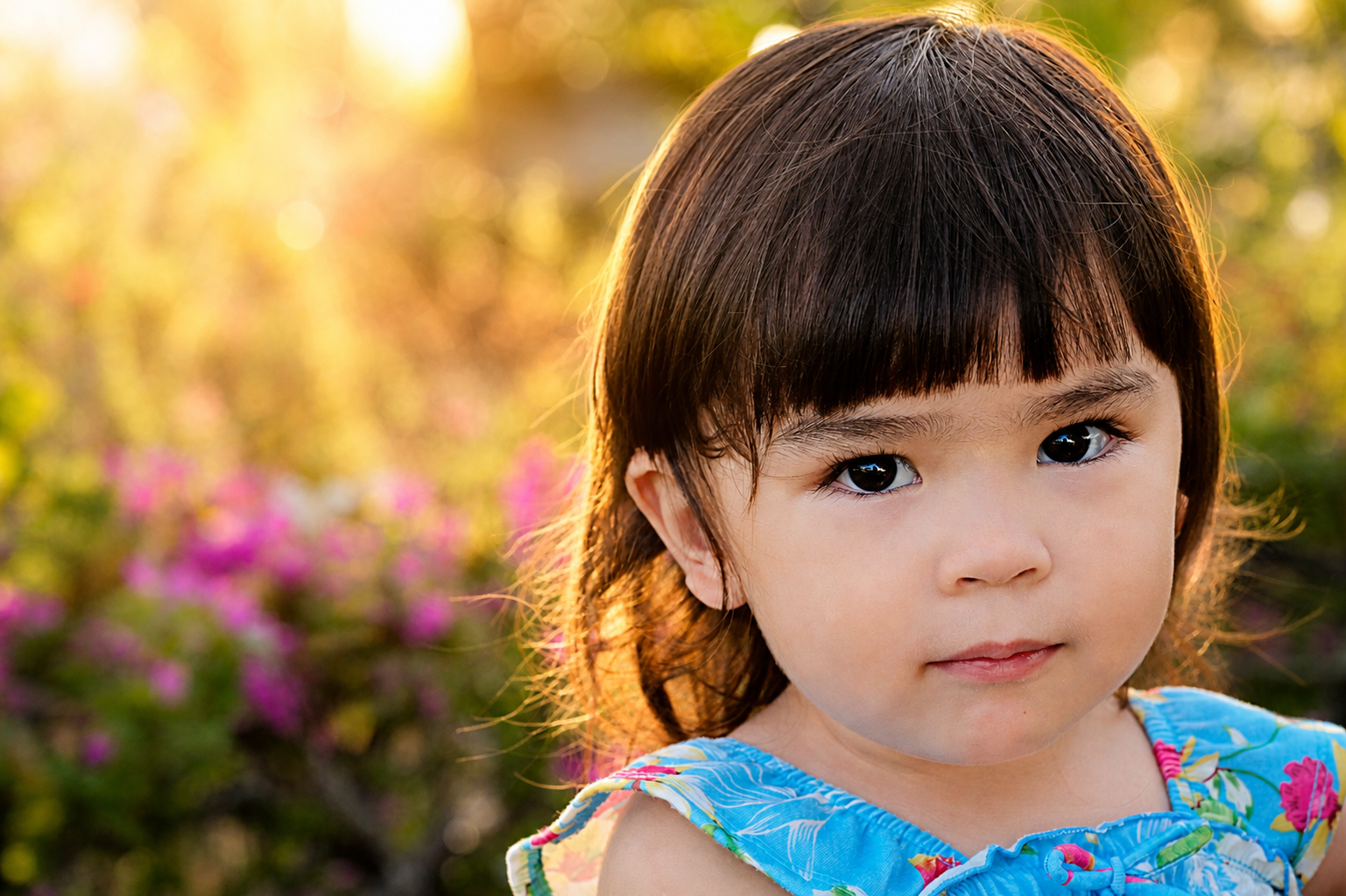 A young girl with brown hair and dark eyes, wearing a blue floral dress, standing outdoors during sunset with blurred pink flowers and green foliage in the background.