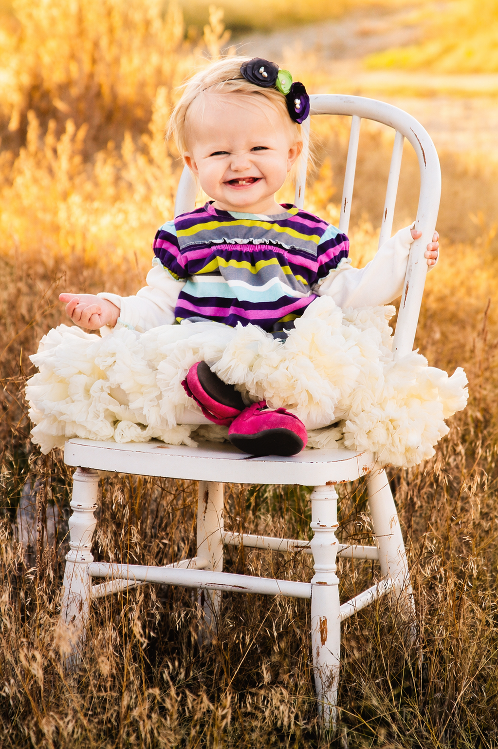 A smiling baby girl with blond hair in a white dress with a purple, green, and black striped top, sitting on a vintage white wooden chair in a field of tall grass during autumn. She is wearing pink shoes and has a black, purple, and green floral head