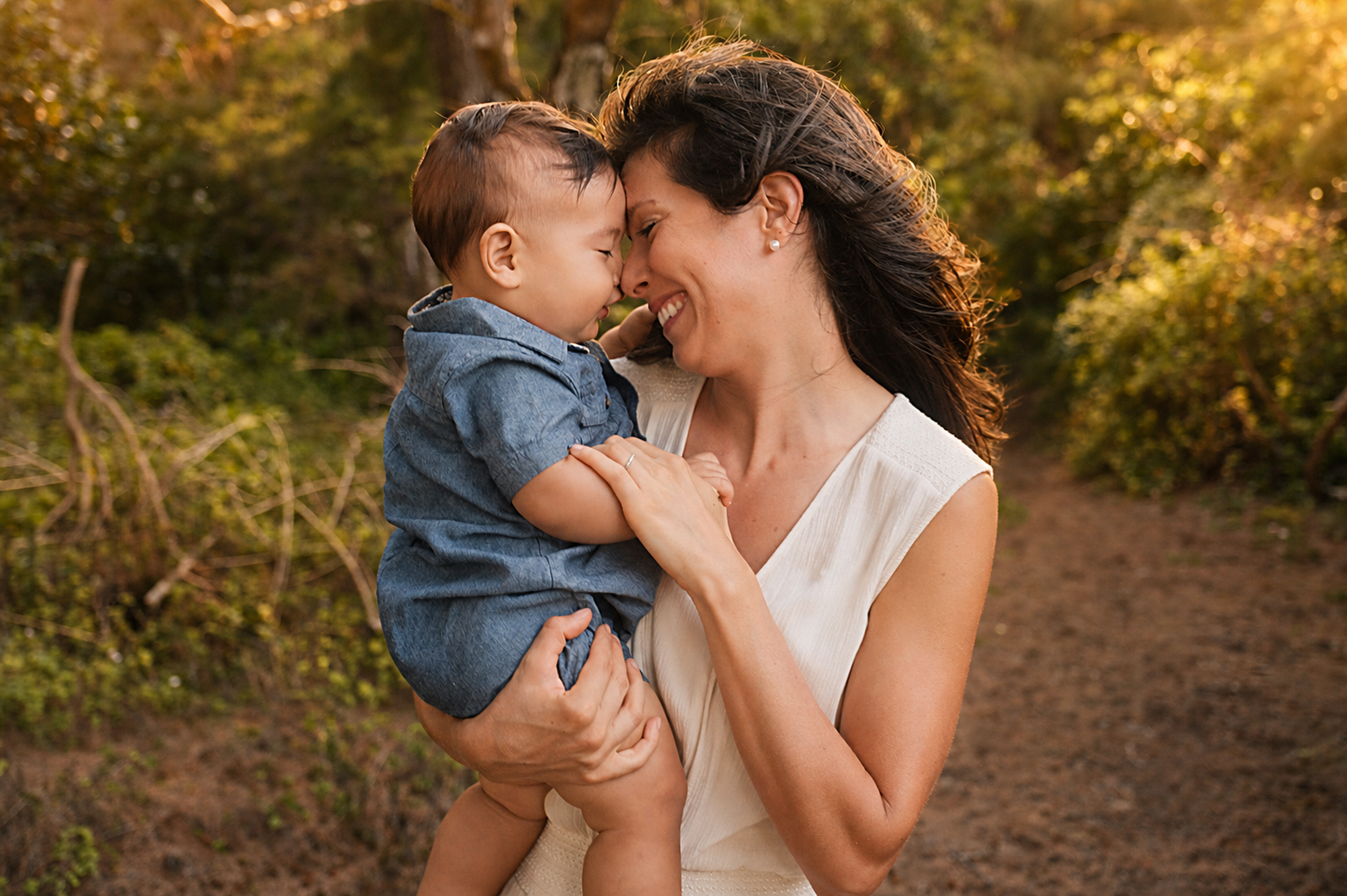 Mom holding a young boy, both touching foreheads, smiling, outdoors in a wooded area during golden hour.