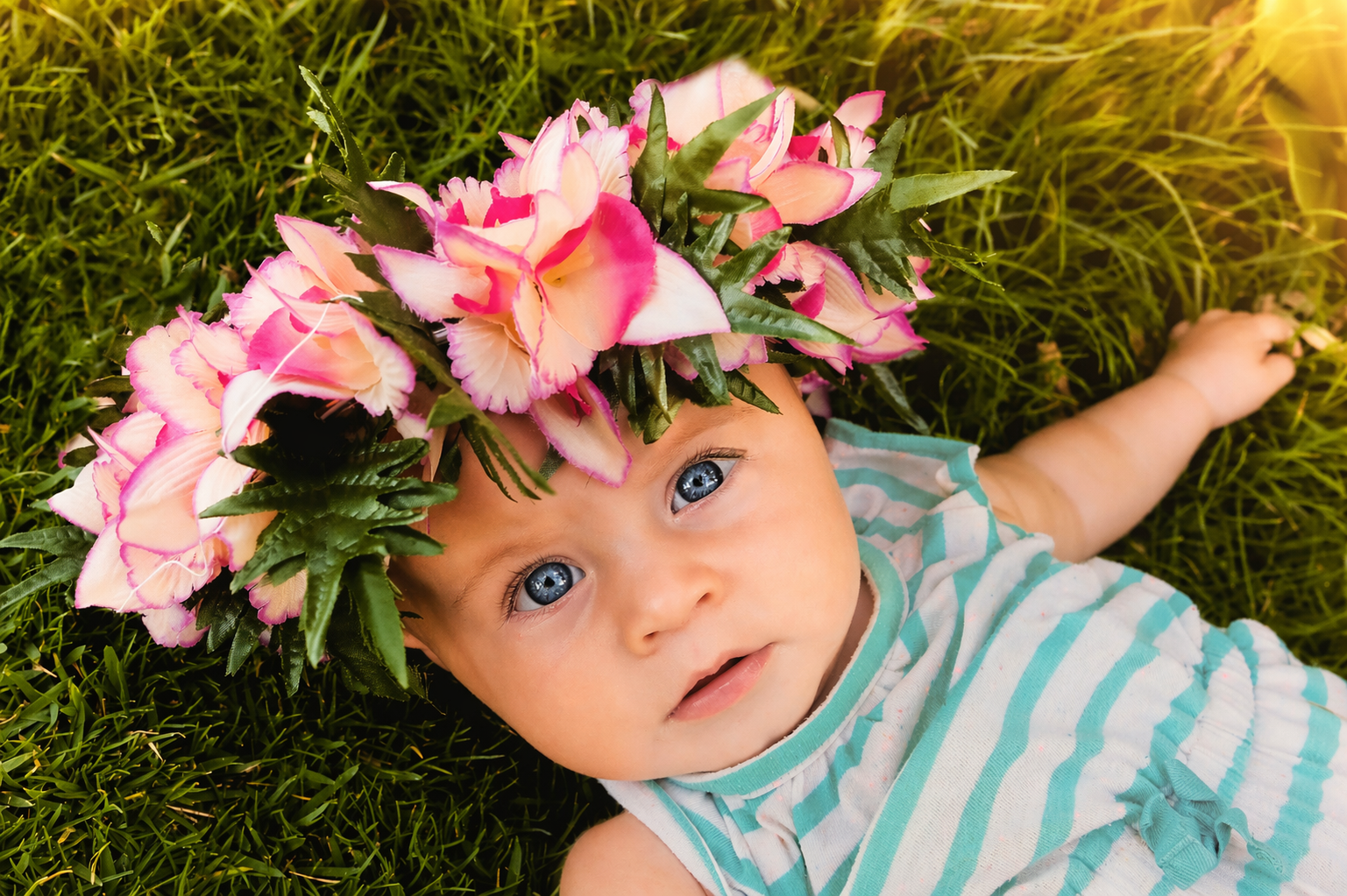 Baby lying on grass wearing a flower crown made of pink and white flowers and green leaves, looking up with blue eyes