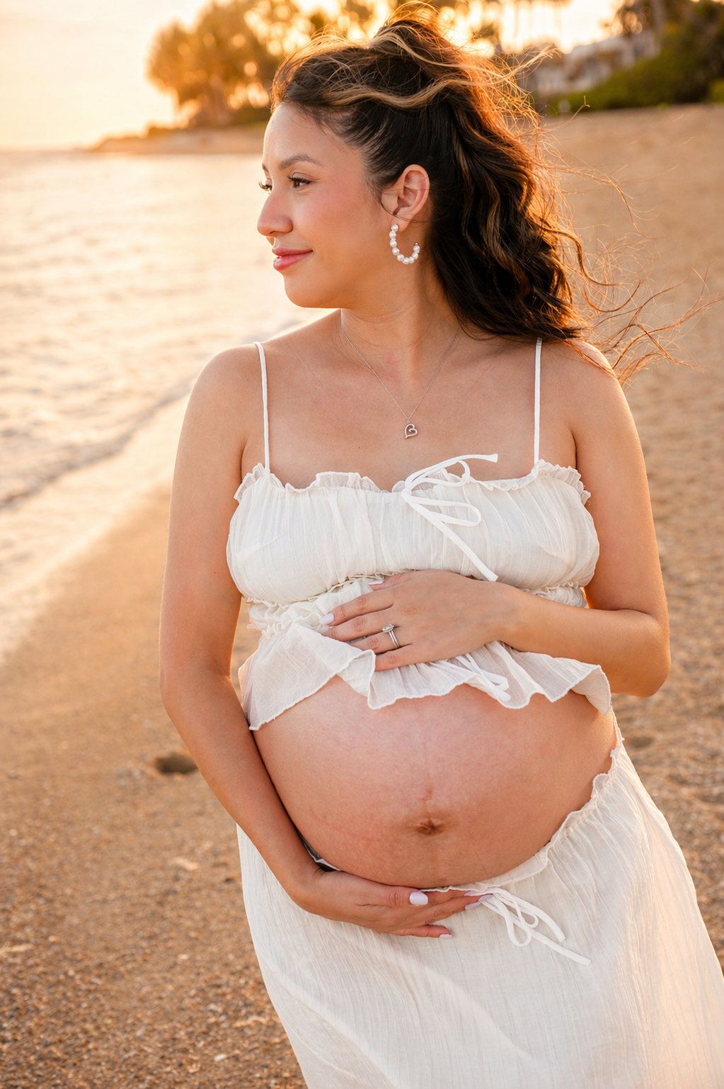 A pregnant woman standing on a beach during sunset, wearing a white dress and jewelry, with her hand on her belly and a serene expression.