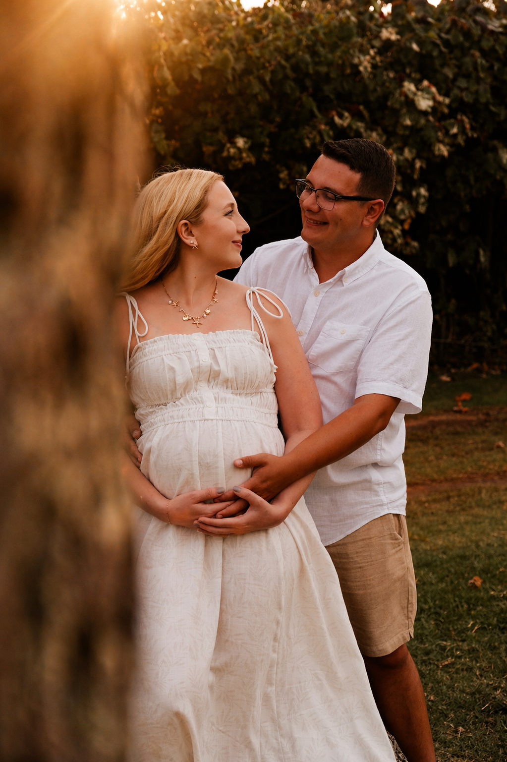 A pregnant woman and a man stand outdoors during sunset, gazing at each other. The woman is wearing a white dress and jewelry, and the man is in a white shirt and shorts. Both are smiling and holding her belly, with trees in the background.