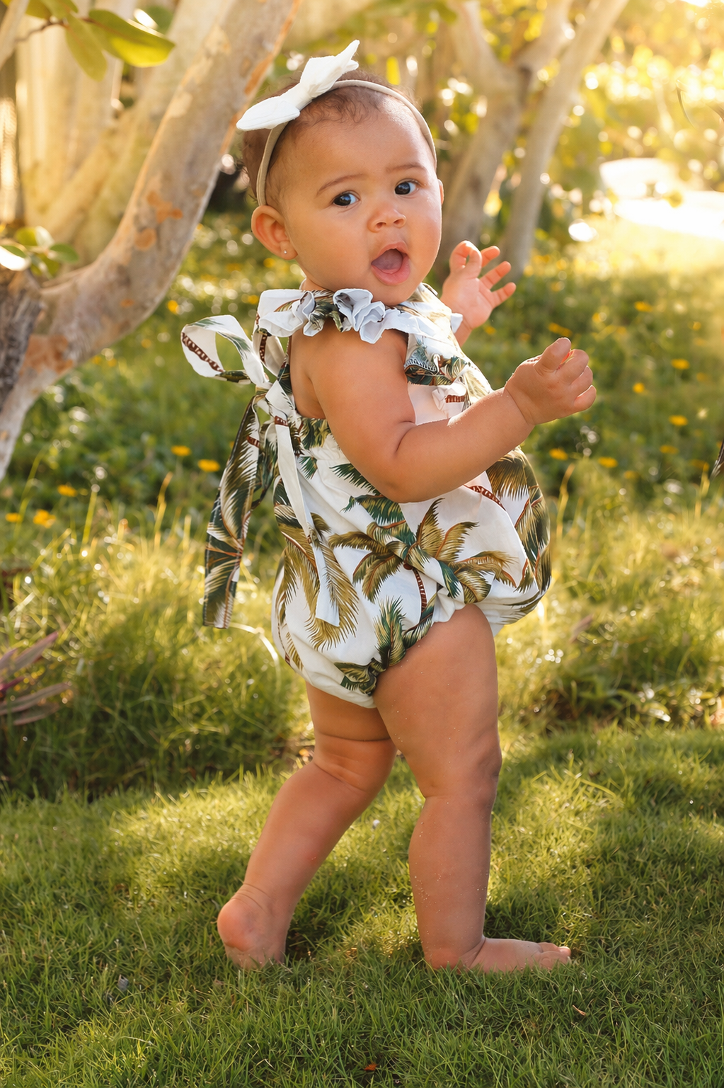 Young child standing outdoors on grassy ground, wearing a tropical print dress with ruffled straps and a matching headband, with a tree and sunlight in the background.