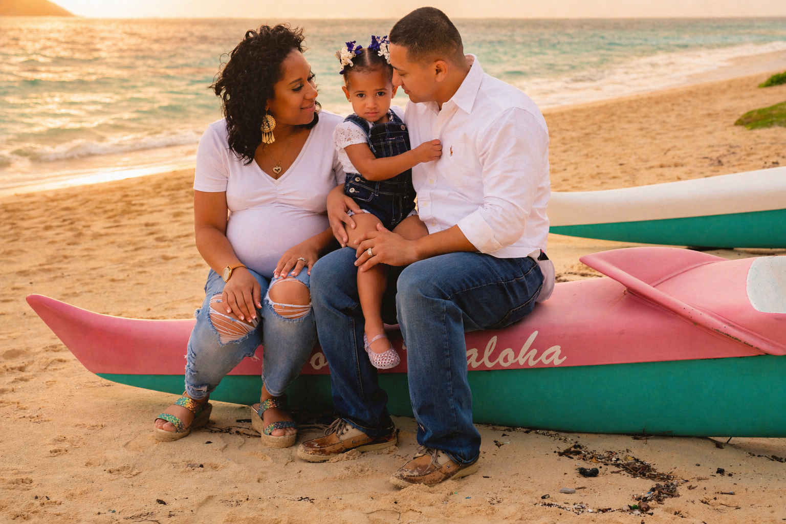 A family of three sitting on pink and teal paddleboards on a sandy beach during sunset, with the ocean in the background. The woman is pregnant, wearing a white t-shirt and ripped jeans, and the man is holding their young daughter, who has hair with 
