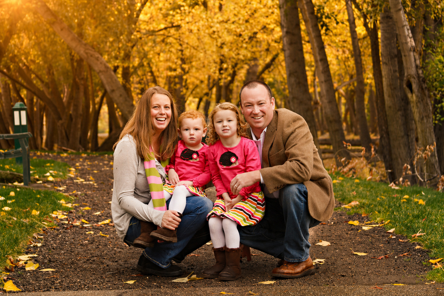 Family of four, including two young girls, smiling and squatting on a forest path during fall. The children are wearing pink dresses with sheep appliqué and striped skirts, while the mother wears a gray sweater with a pink and green striped scarf, an