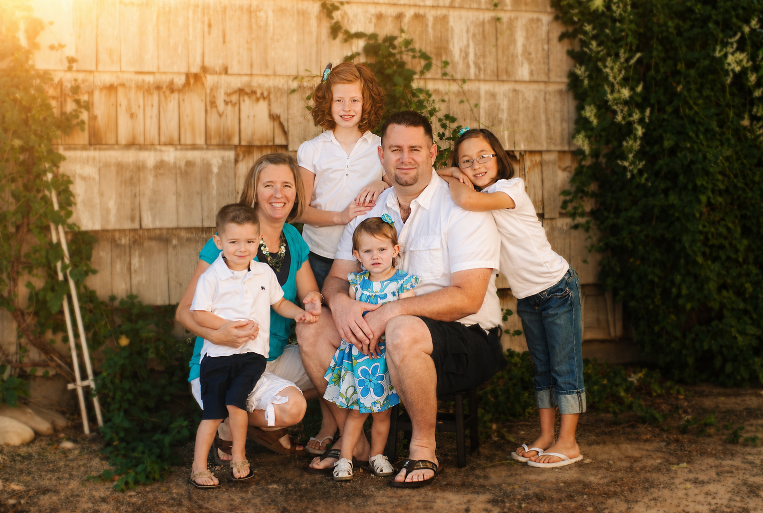 A family of six, including two adults and four children, posing outdoors against a wooden fence with greenery, during sunset.