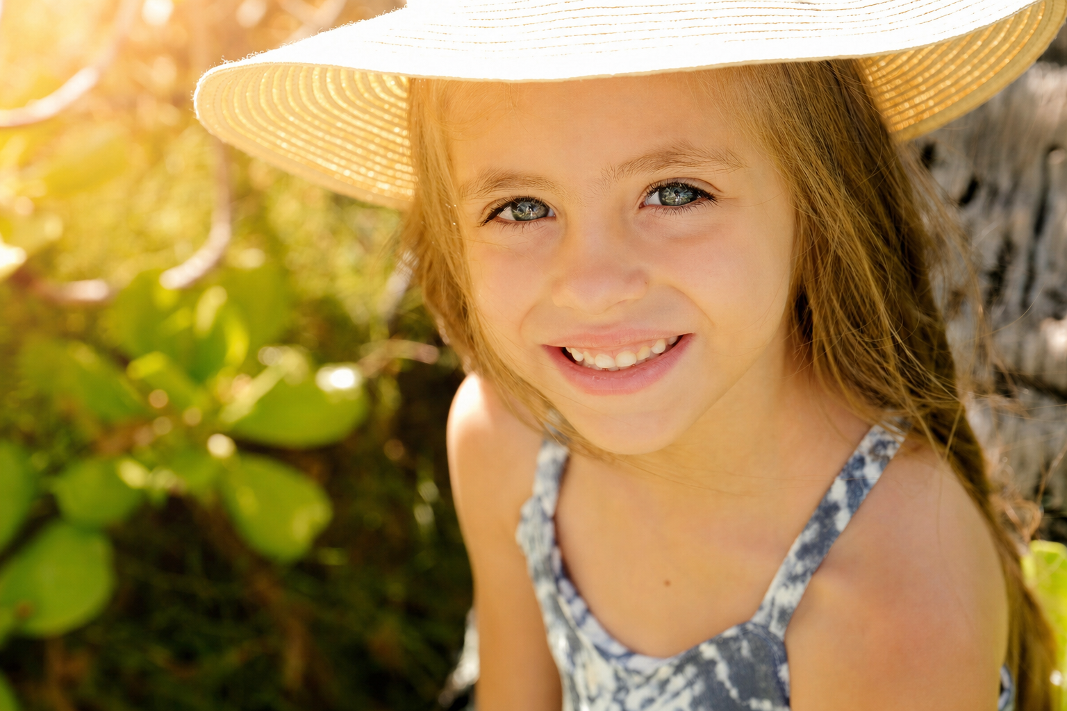 A young girl with blue eyes and long brown hair wearing a straw hat and a blue floral dress, smiling outdoors in sunlight.