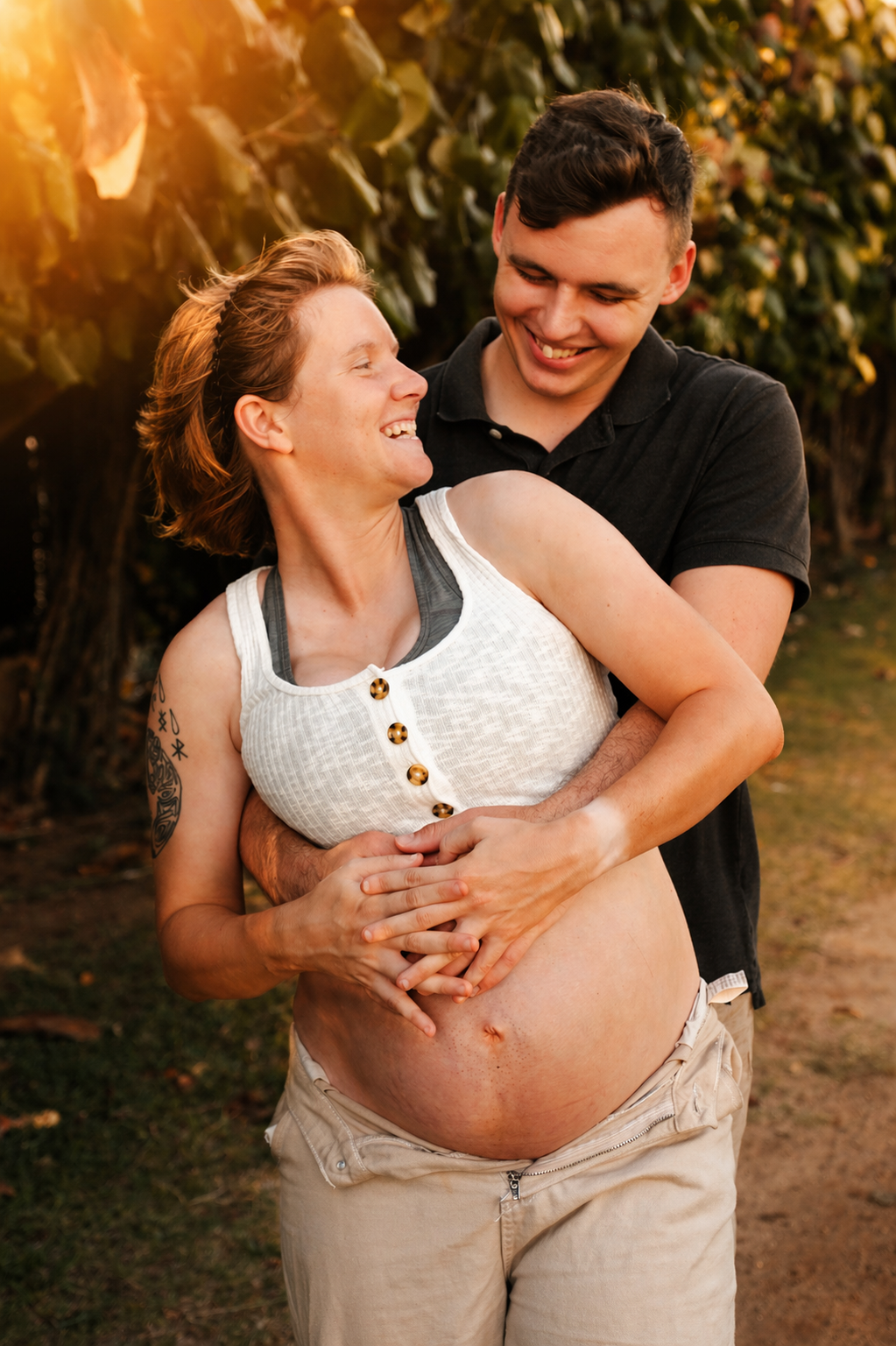 A couple sharing a joyful moment outdoors during sunset, with the woman showing her pregnant belly and both smiling and looking happy.
