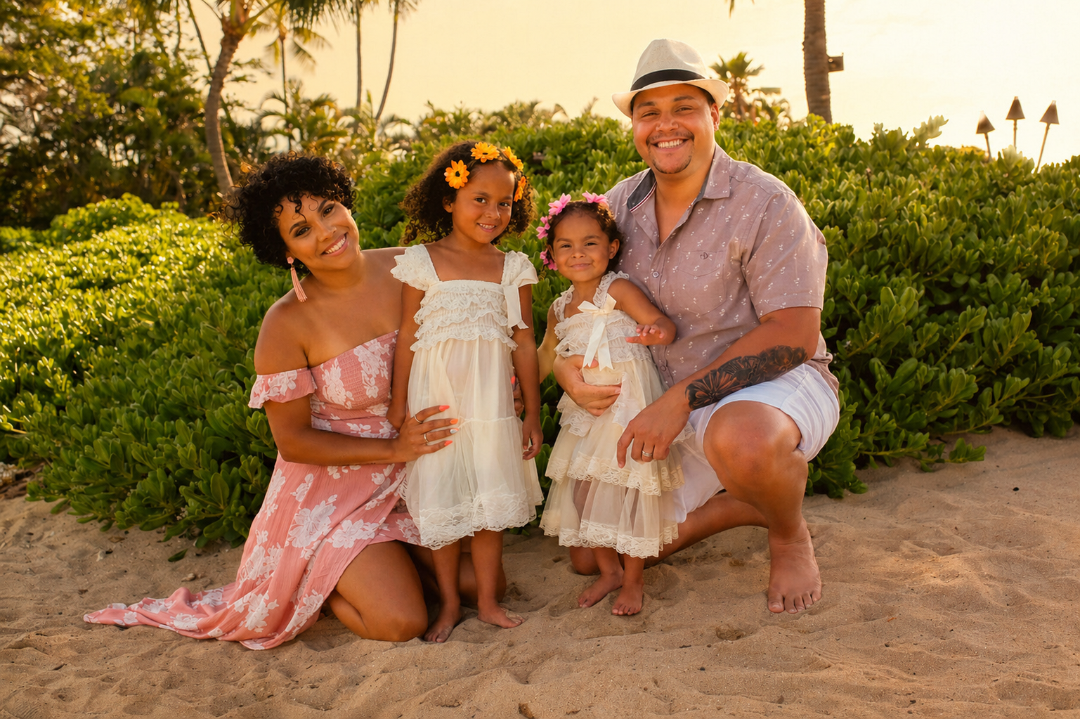 A family of four smiling and posing on a sandy beach during sunset, surrounded by green bushes and tall palm trees. The mother is kneeling on the left, wearing a pink floral dress. The father is kneeling on the right, wearing a light-colored shirt, s
