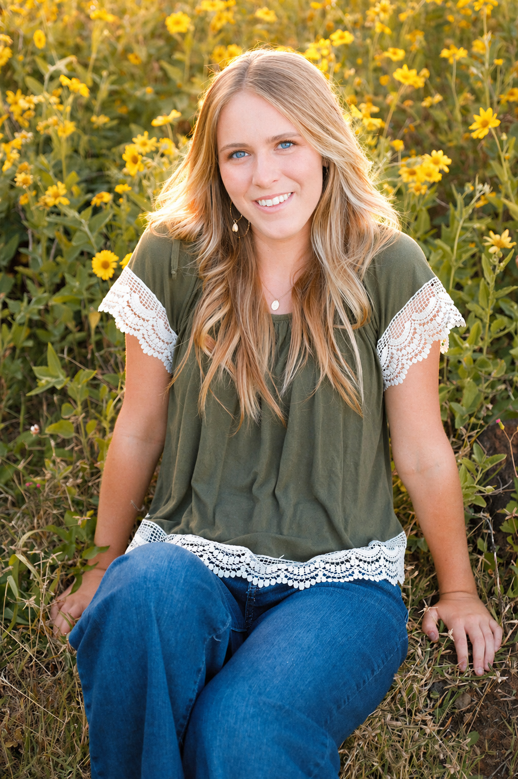 A young woman with long blonde hair and blue eyes sitting on the grass among yellow flowers, smiling at the camera during sunset.