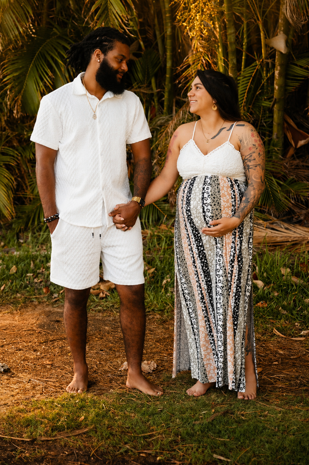 A couple holding hands outdoors, woman is pregnant, standing on grass with tropical plants behind them, warm sunset lighting.