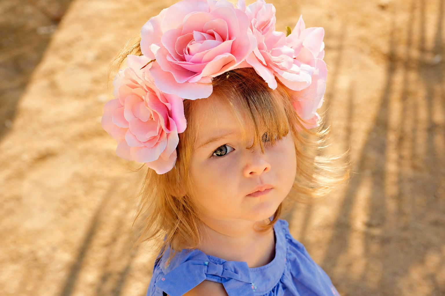 A young girl with red hair wearing a large pink flower crown and a blue dress outdoors.