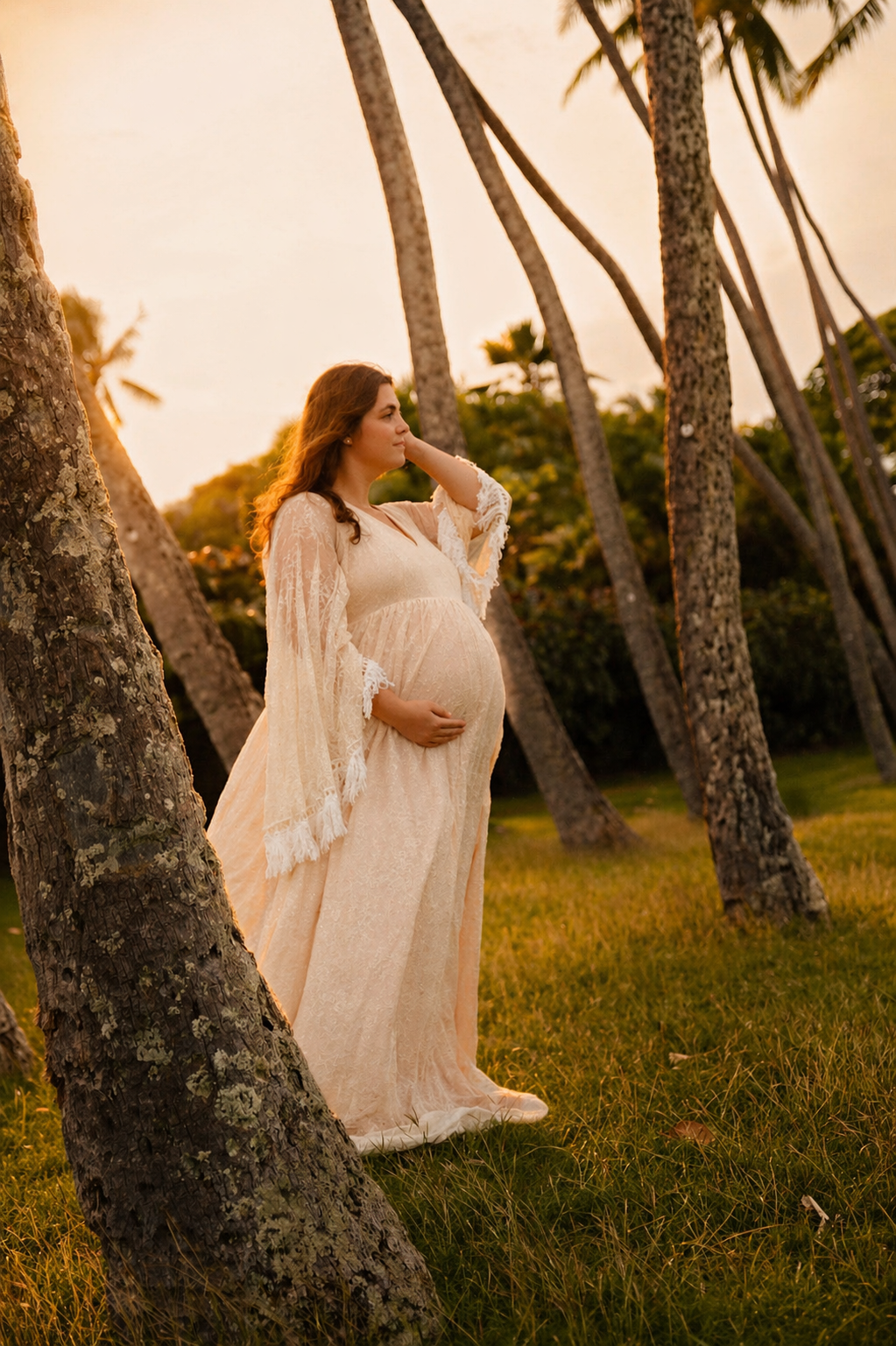 A pregnant woman in a flowing beige lace dress stands among palm trees during sunset, gently touching her belly and looking into the distance.