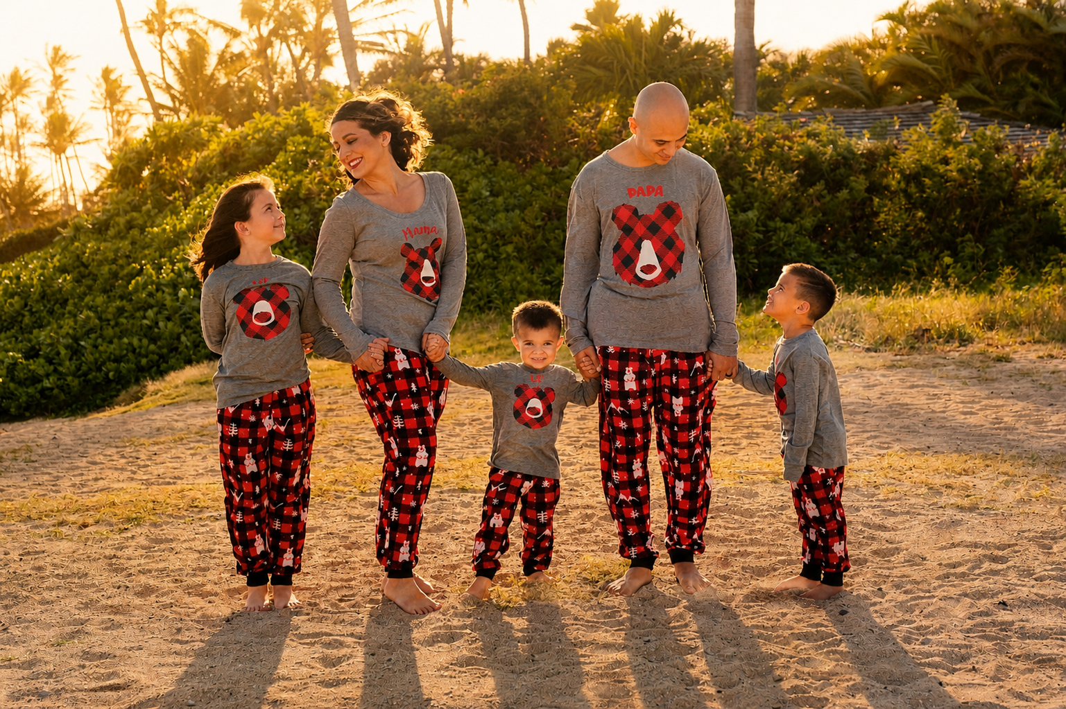 Family of six on a beach at sunset, wearing matching gray holiday pajamas with red plaid bear designs and pajamas with snowmen, holding hands and smiling.