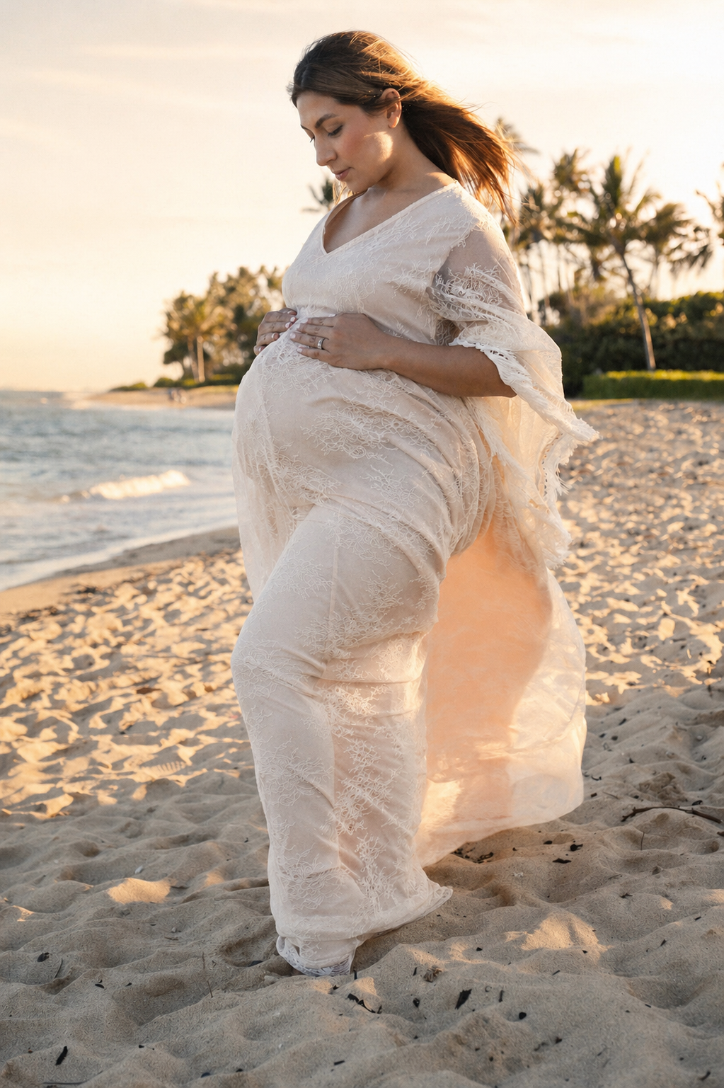 A pregnant woman wearing a light-colored lace dress standing on a sandy beach at sunset, holding her belly with both hands and looking down.