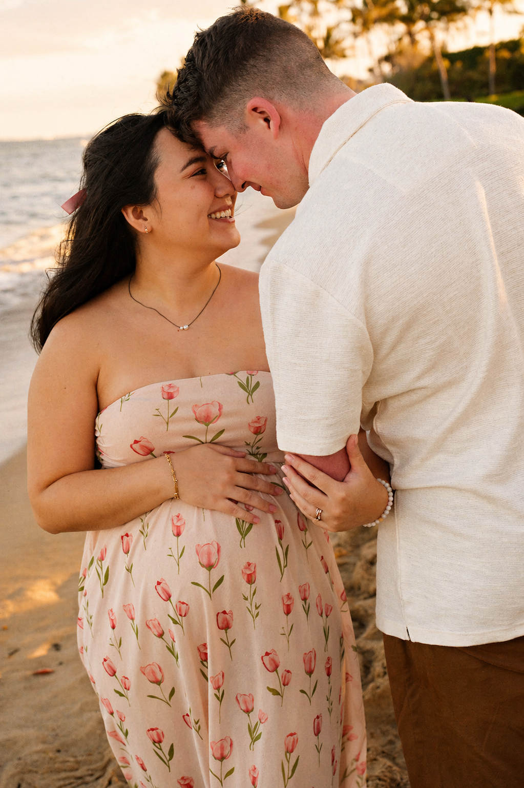 A couple at the beach during sunset, holding hands and touching foreheads, smiling at each other, with the ocean and shoreline in the background.