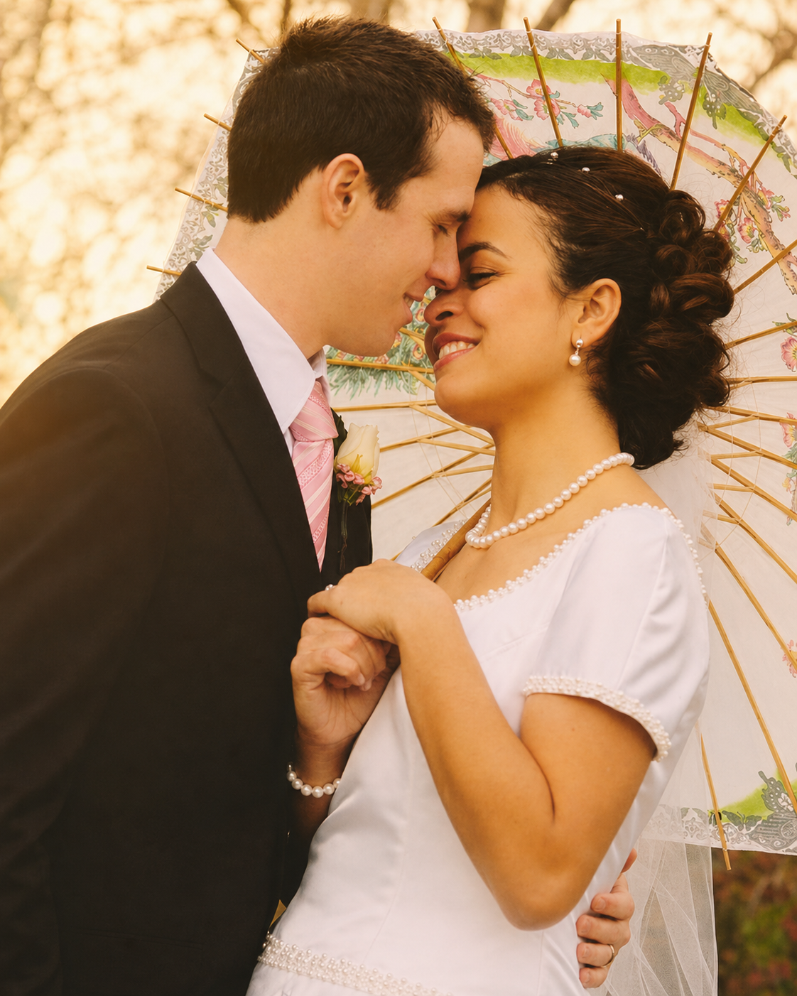 A bride and groom sharing a romantic moment under a colorful parasol, with their foreheads touching and eyes closed, at a wedding celebration.
