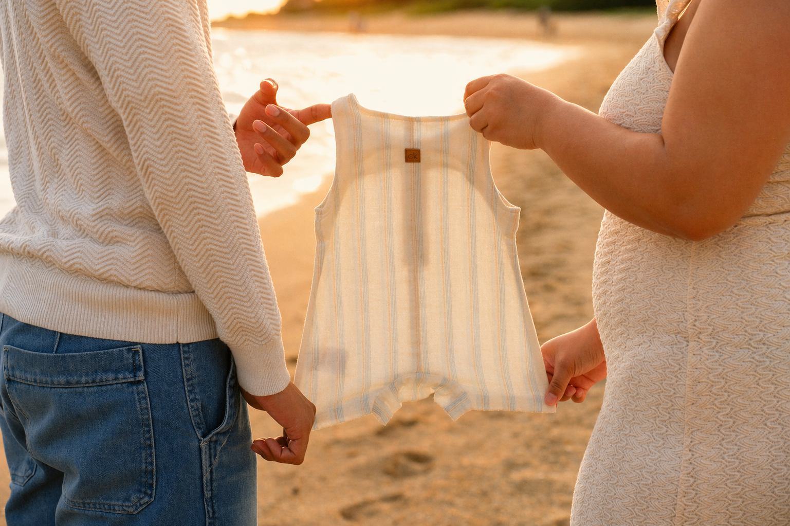 Two people holding up a small striped baby romper on a beach during sunset.