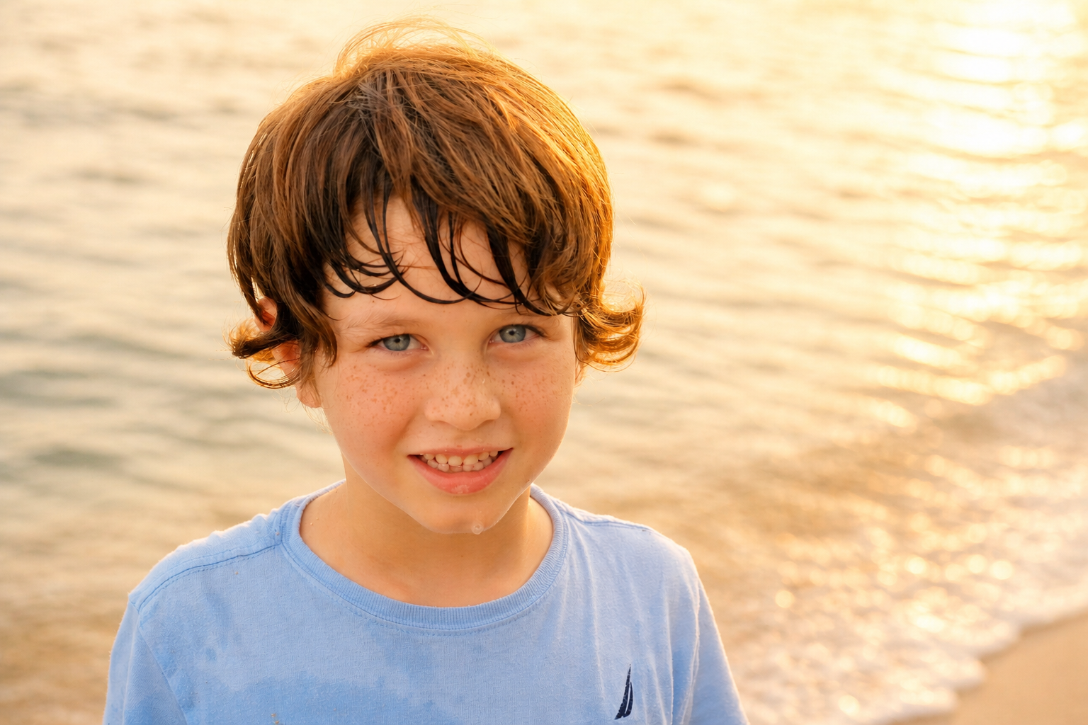A young boy with wet, brown hair and blue eyes smiling at the camera at the beach during sunset.