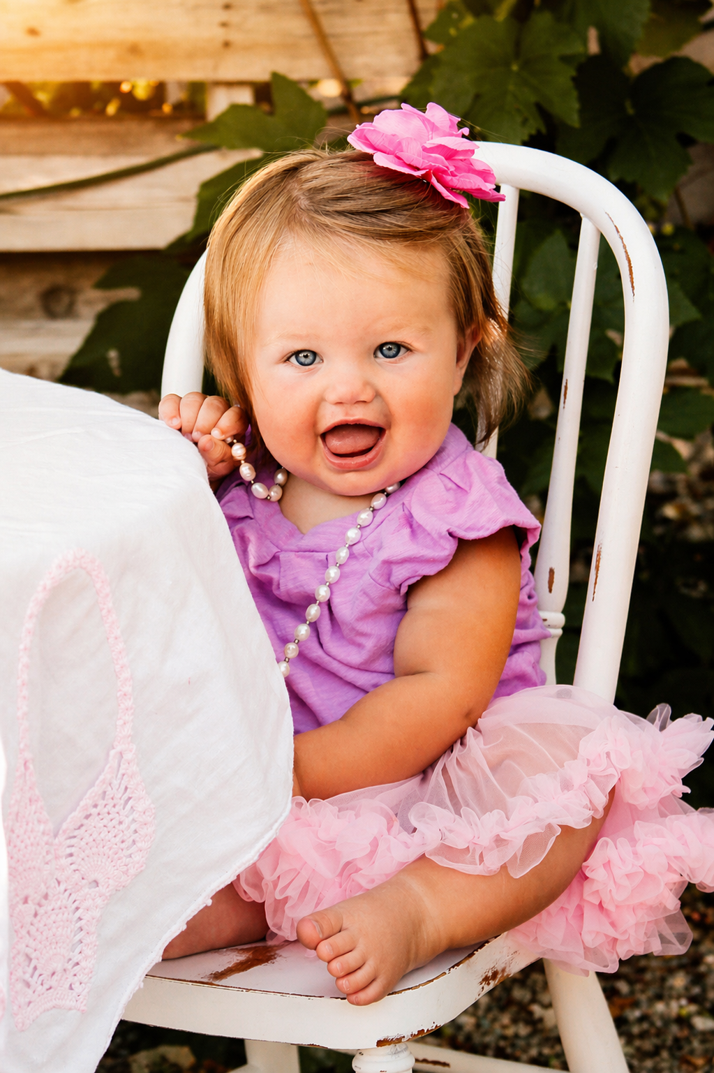 A young girl with light brown hair, blue eyes, and fair skin, wearing a purple dress with ruffled sleeves, a pink tutu, a pearl necklace, and a large pink flower hair accessory, sitting on a white distressed wooden chair outdoors, with green foliage 