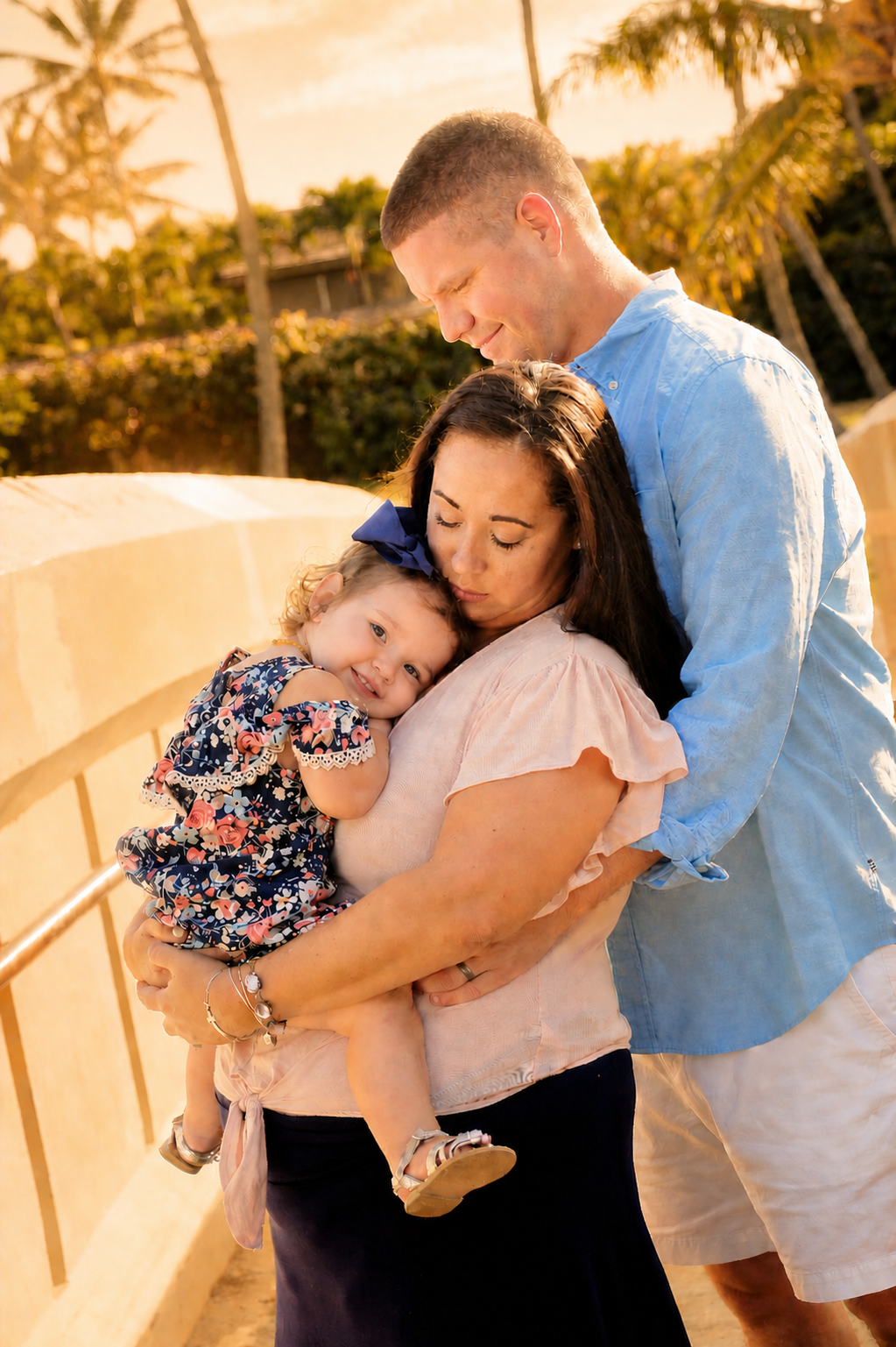 A family of three, a man, a woman, and a young girl, are hugging outdoors during sunset. The little girl is smiling and resting her head on the woman's shoulder, while the woman gently holds her. The man leans in affectionately, and they are surround