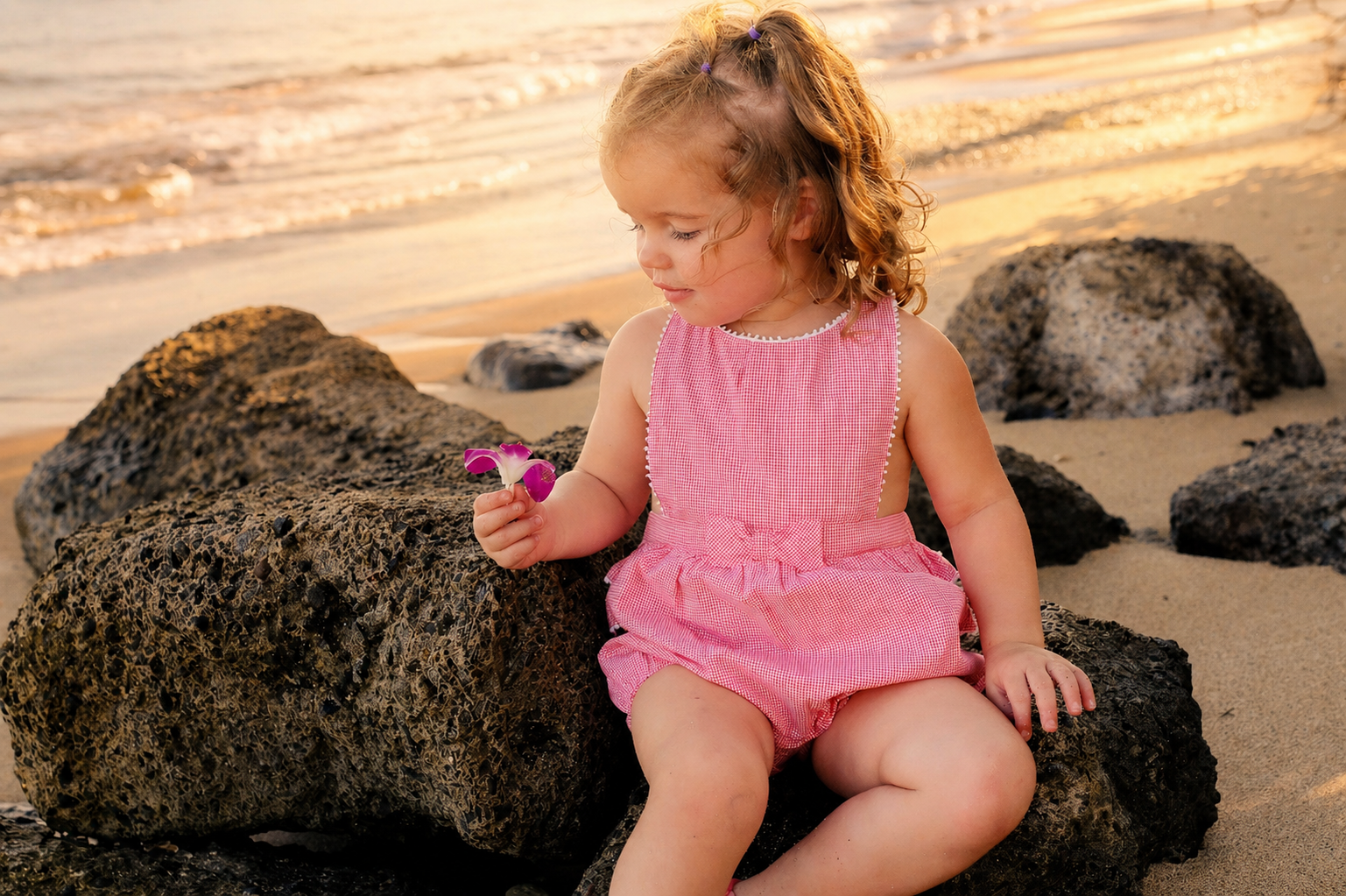 A young girl with curly hair sitting on a rock at the beach, holding a pink flower, with sand and rocks around her and the ocean in the background.