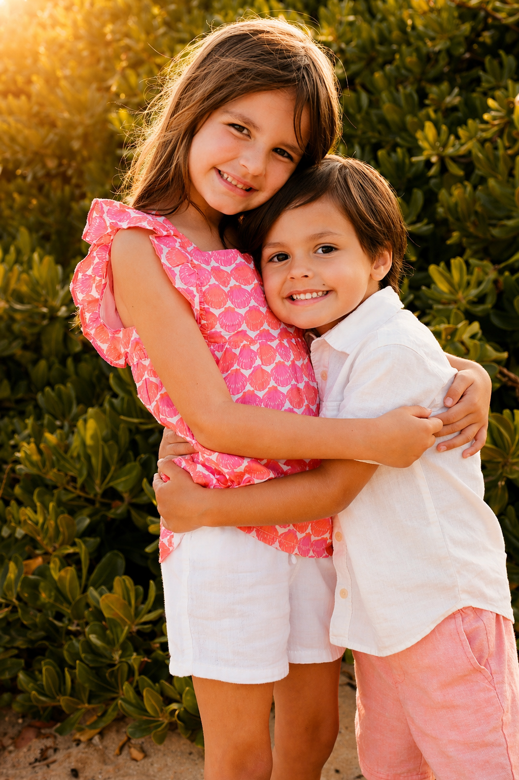 Two children, a girl and a boy, hugging outdoors during sunset, smiling, surrounded by green bushes.