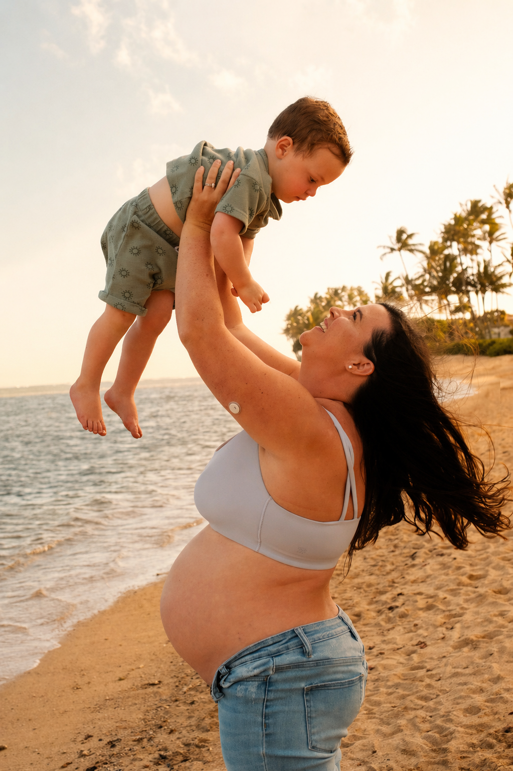 A woman with a pregnant belly holding a young boy up in the air on a beach during sunset.