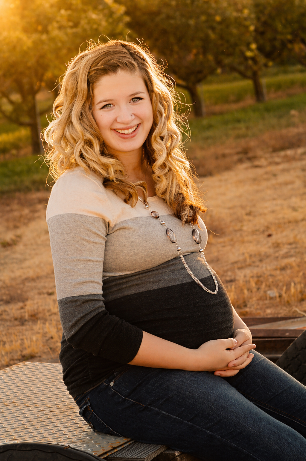 A pregnant woman with curly blonde hair smiling outdoors during sunset, sitting on a metal surface, wearing a color-blocked long sleeve top and a necklace, with trees in the background.