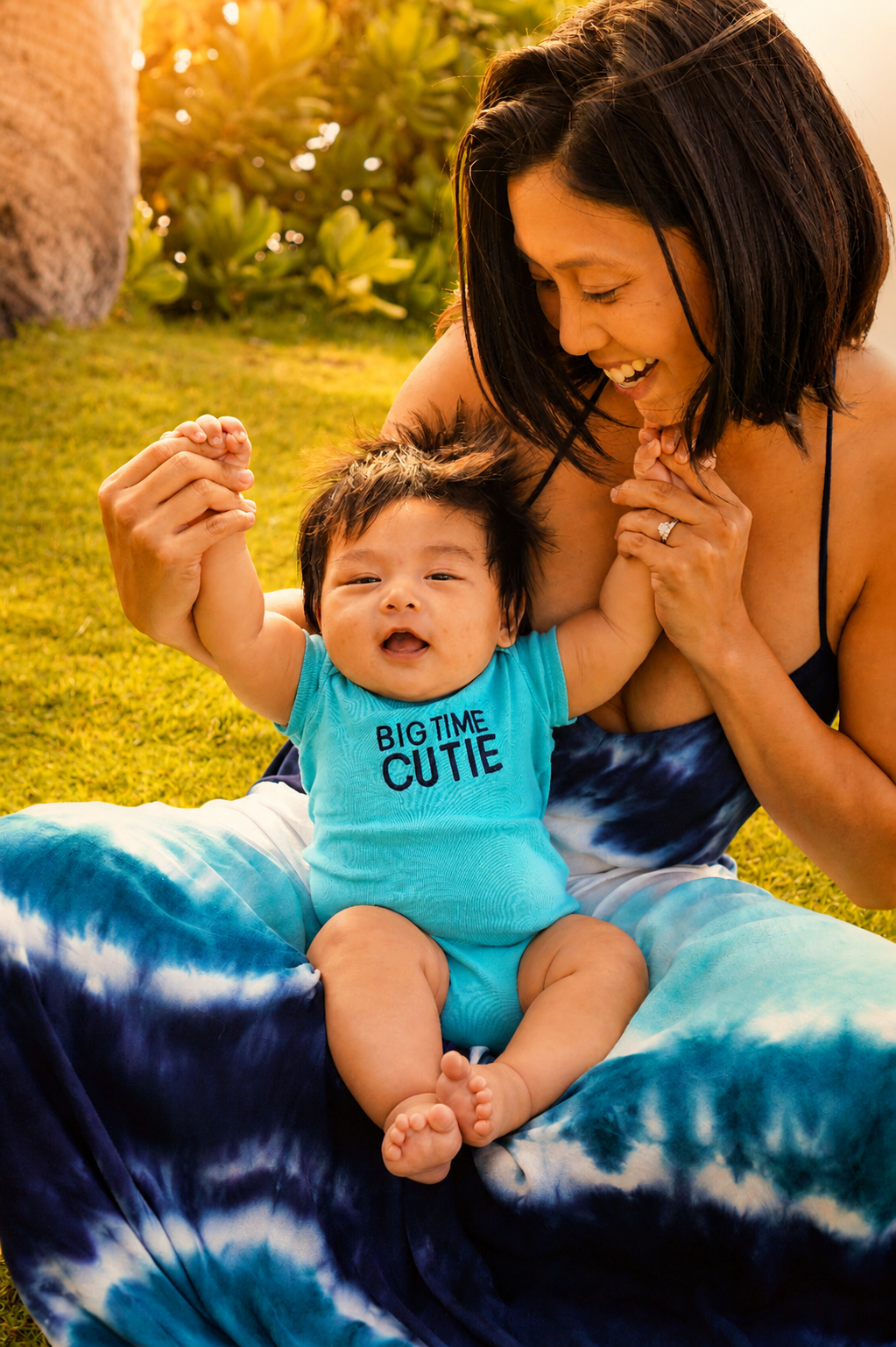 A woman and a baby boy sitting on the grass outdoors during sunset, holding hands. The woman is smiling and wearing a black and blue tie-dye dress, while the baby is wearing a blue shirt that says 'Big Time Cutie' and has an excited expression.