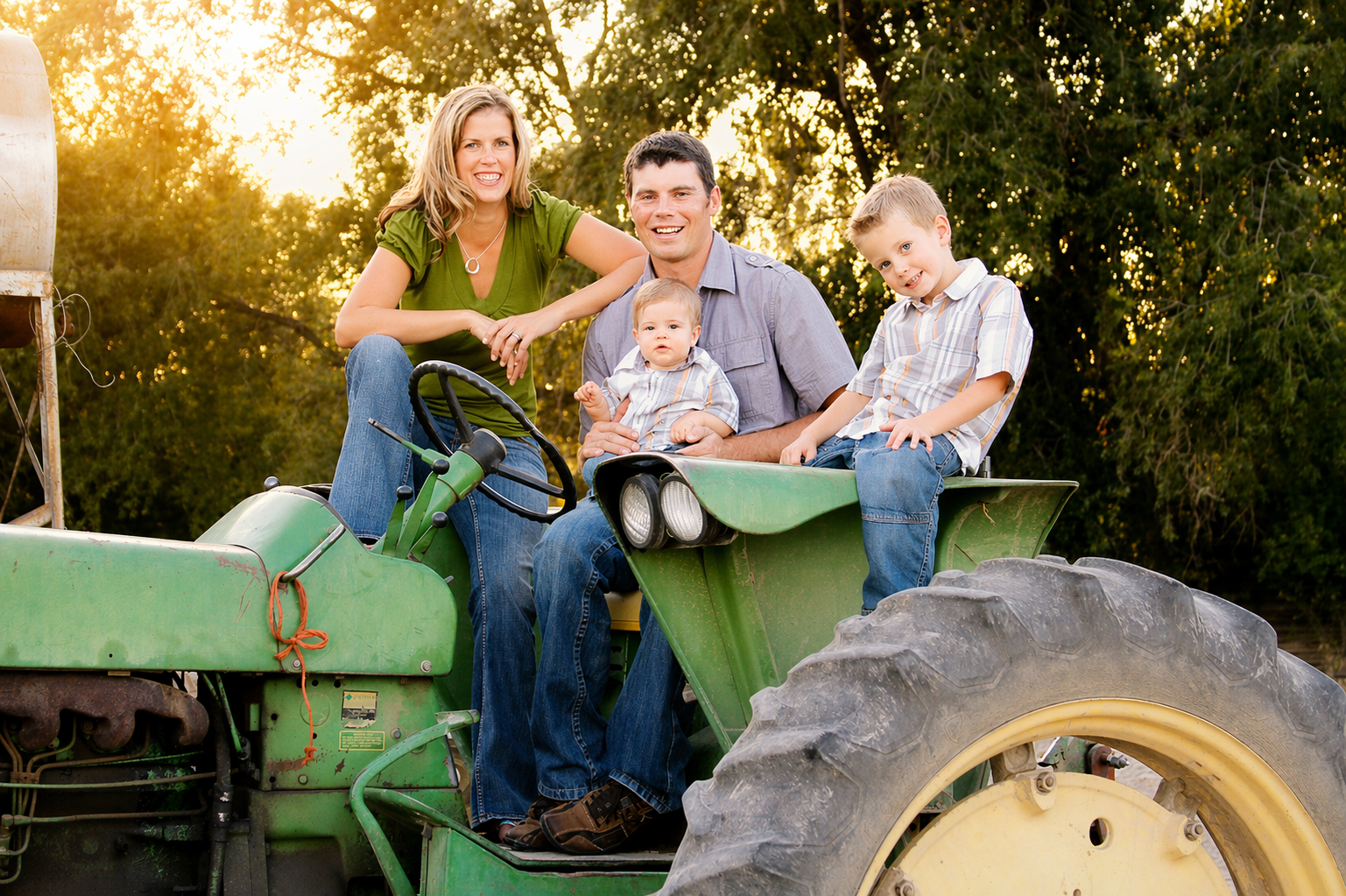 A family of four sitting on a green tractor in a farm setting during sunset. The family includes a woman in a green shirt, a man in a gray shirt, a young boy, and a baby. They are smiling and enjoying the outdoors with trees in the background.