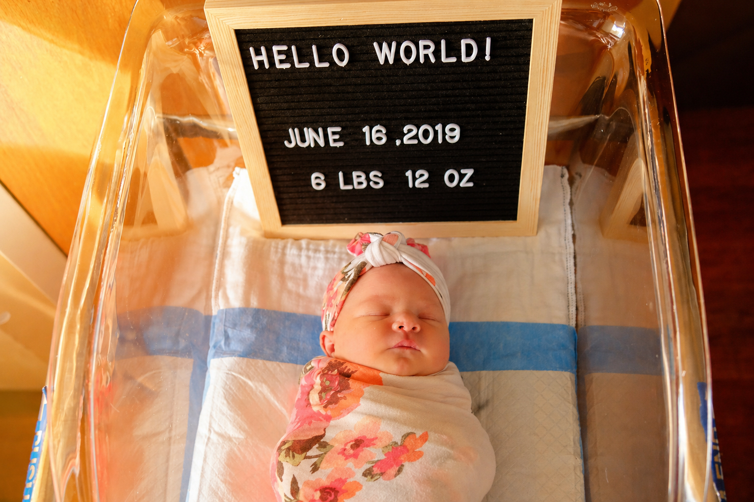 A newborn baby sleeping in a hospital bassinet wrapped in a floral blanket, with a small black letter board displaying the date June 16, 2019, weight 6 lbs 12 oz, and the message 'HELLO WORLD!'