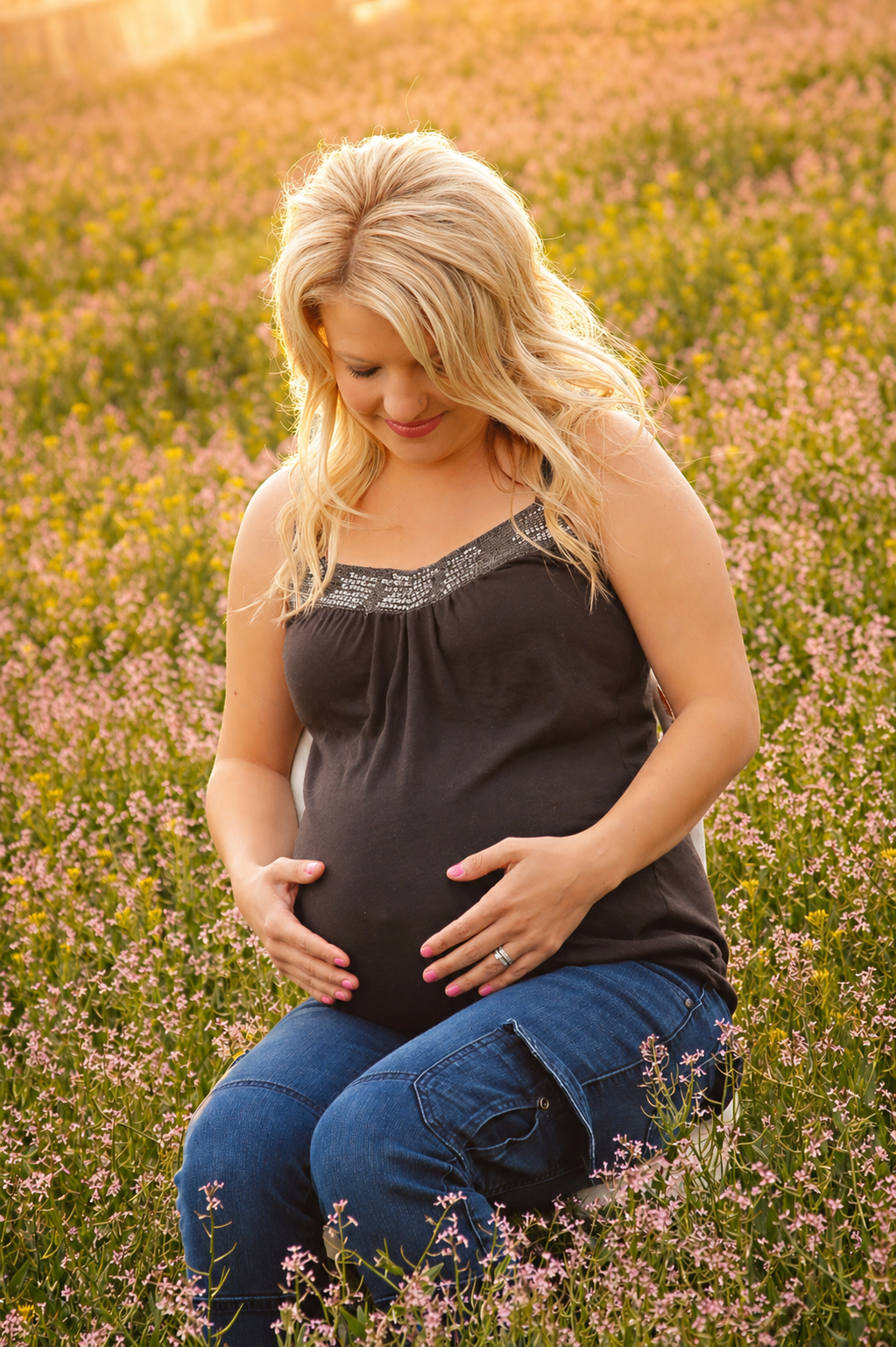 Pregnant woman with blonde hair in a black tank top and jeans kneeling in a field of pink flowers during sunset.
